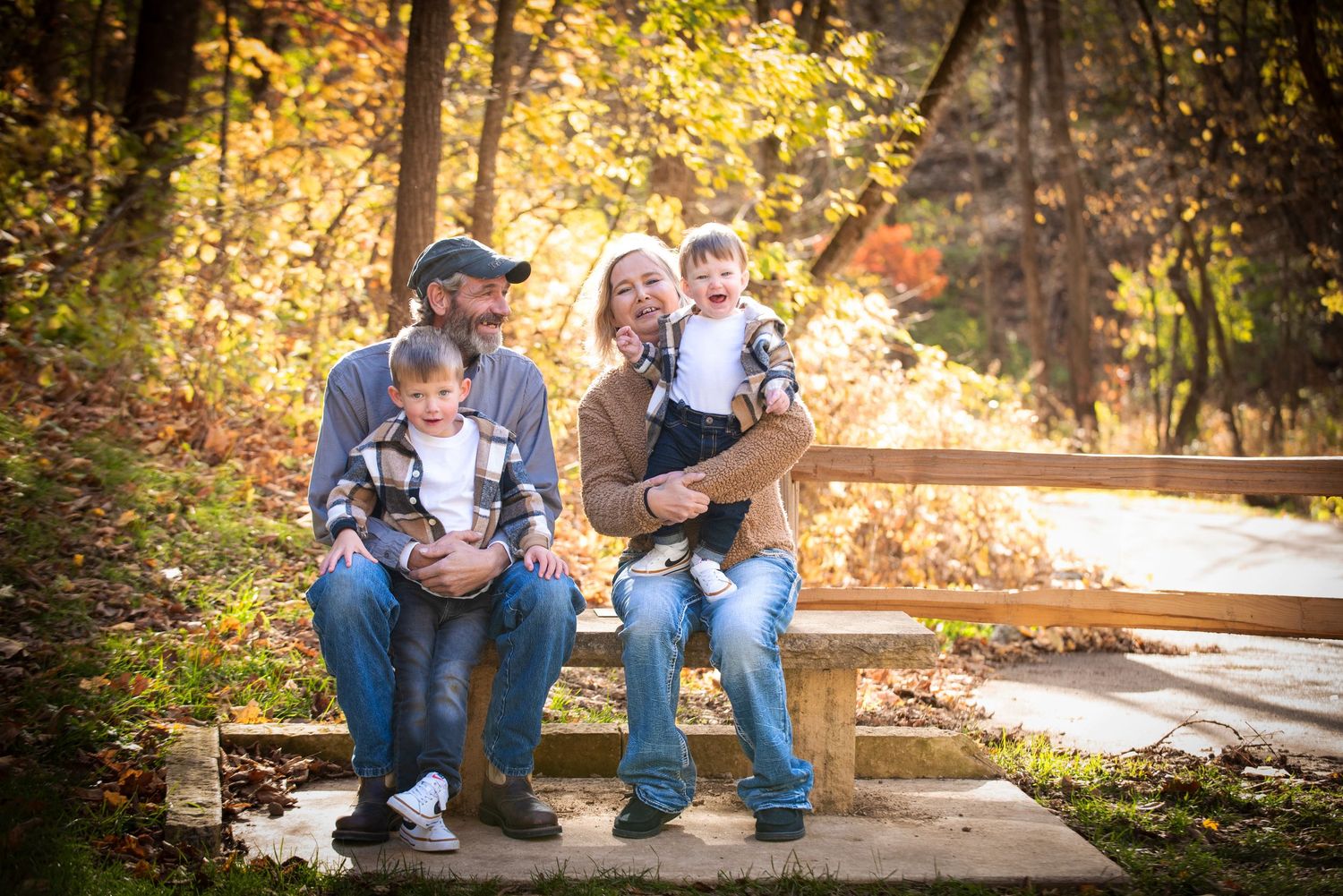 Young couple sits on a wooden bench holding two toddlers surrounded by colorful fall foliage by a stream.
