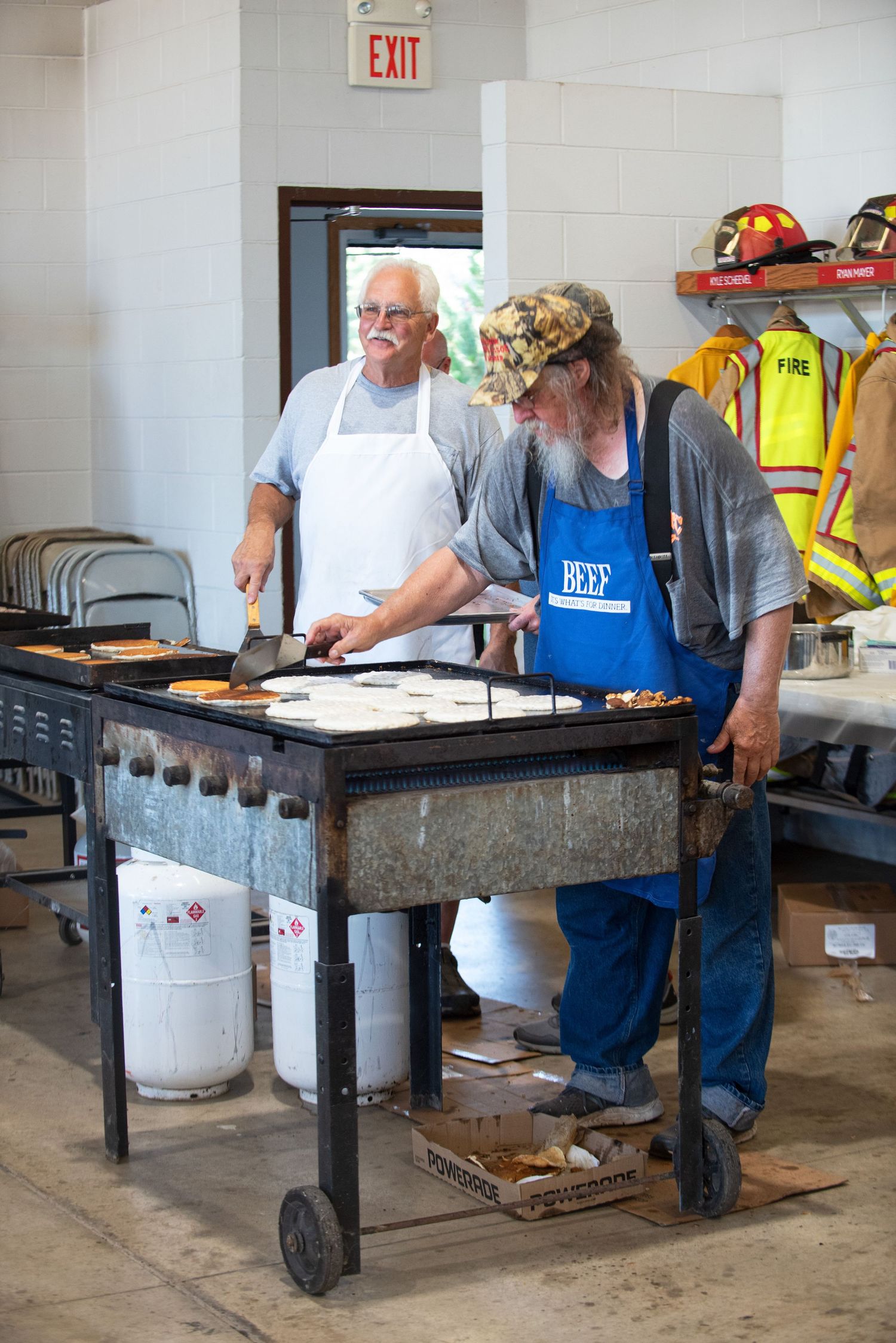 Two men in aprons cook pancakes on a large flat-top griddle at a community breakfast event. 4th of July Celebration in Harmony MN.
