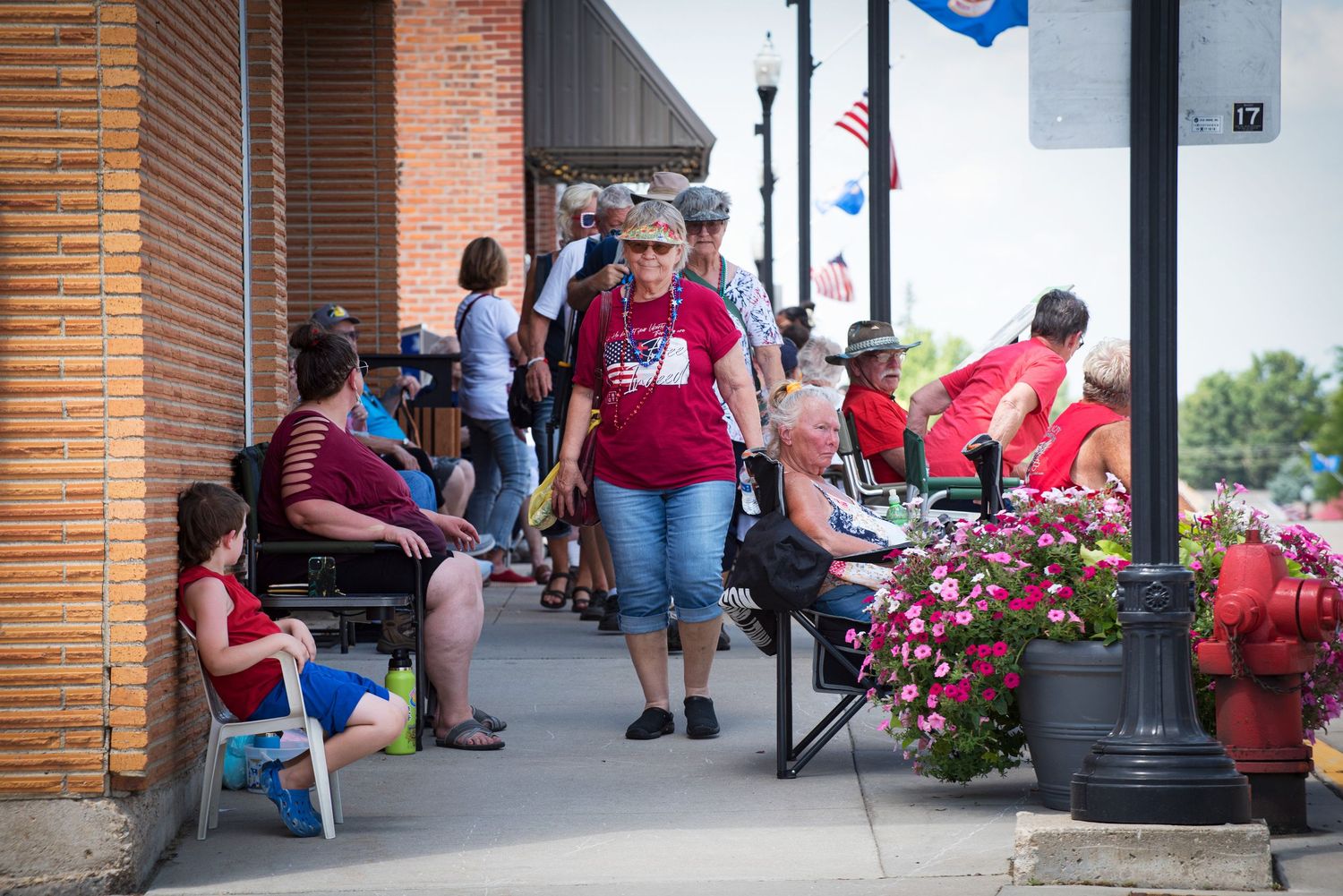 Crowds gather on a brick sidewalk decorated with flowers during a festive small-town summer event. At Harmony MN 4th of July Celebration.