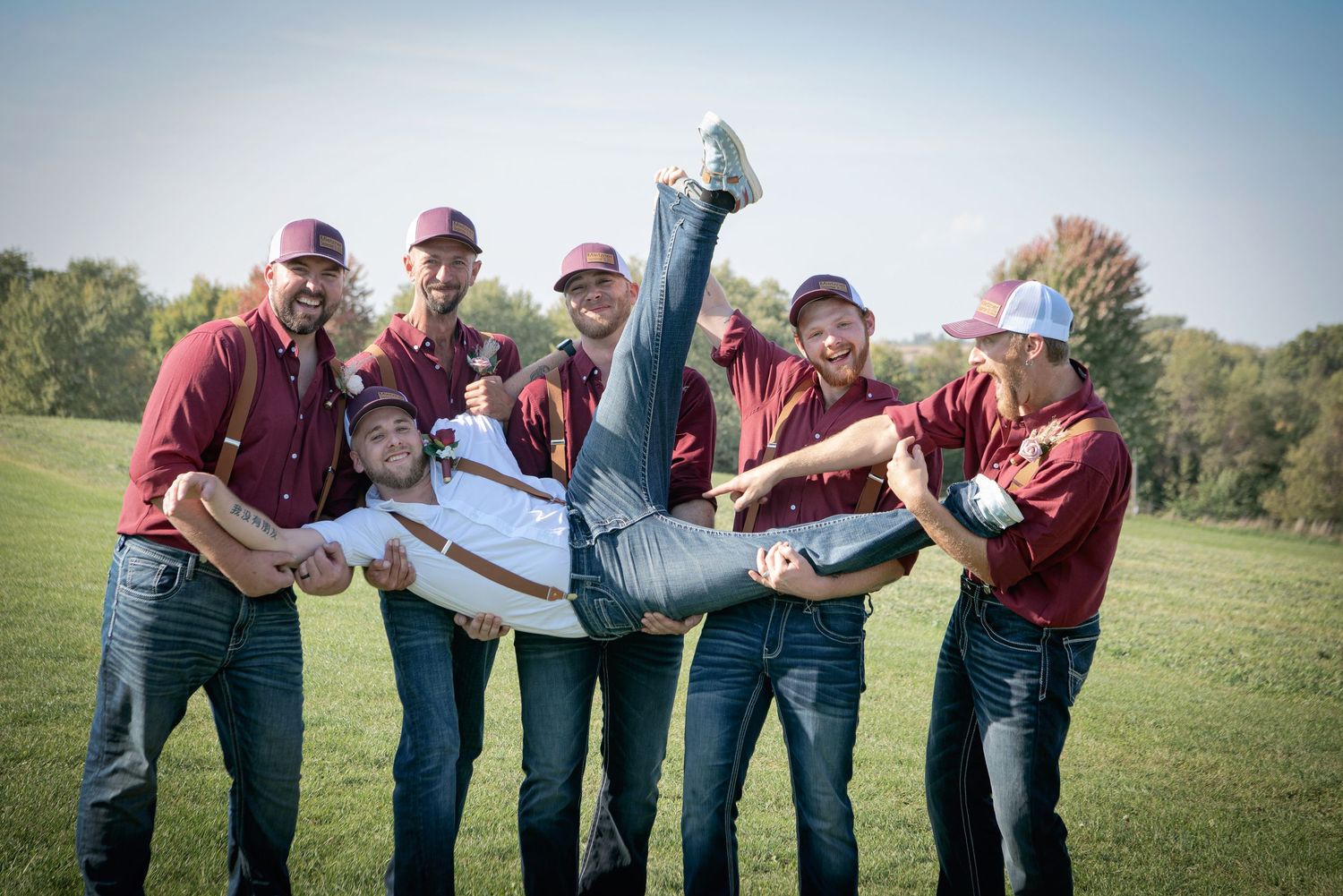 Groomsmen in maroon shirts playfully lift groom horizontally in an outdoor field on wedding day.