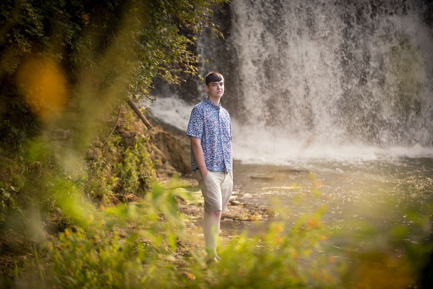 Young man in floral shirt standing near a waterfall framed by lush green foliage.