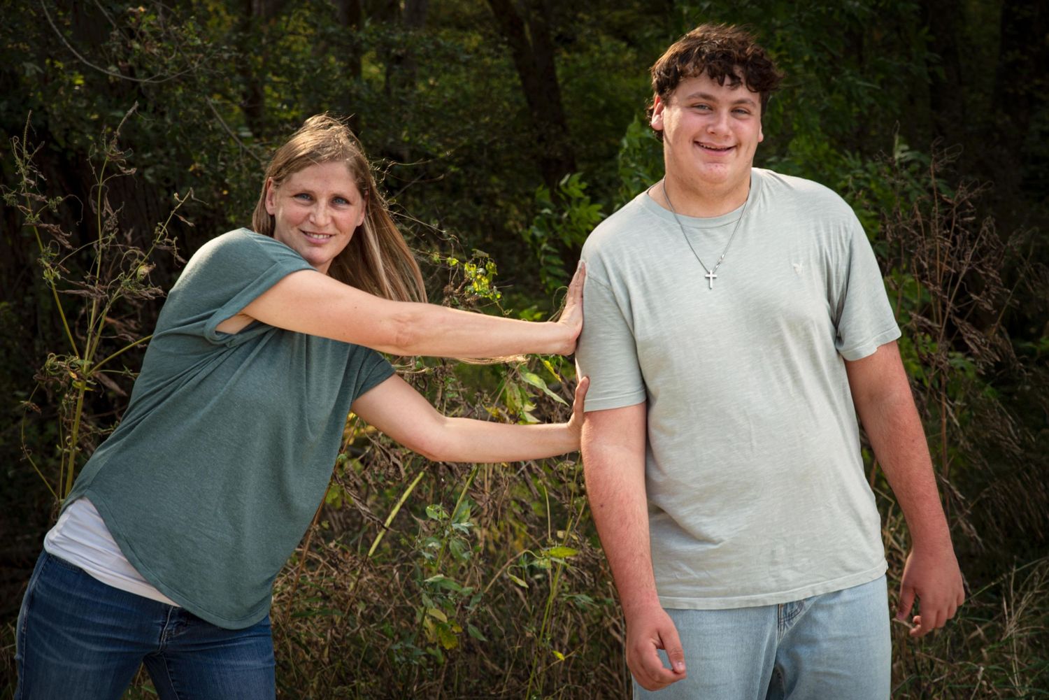 A smiling teenage girl playfully pushes a teenage boy from behind in a wooded area.