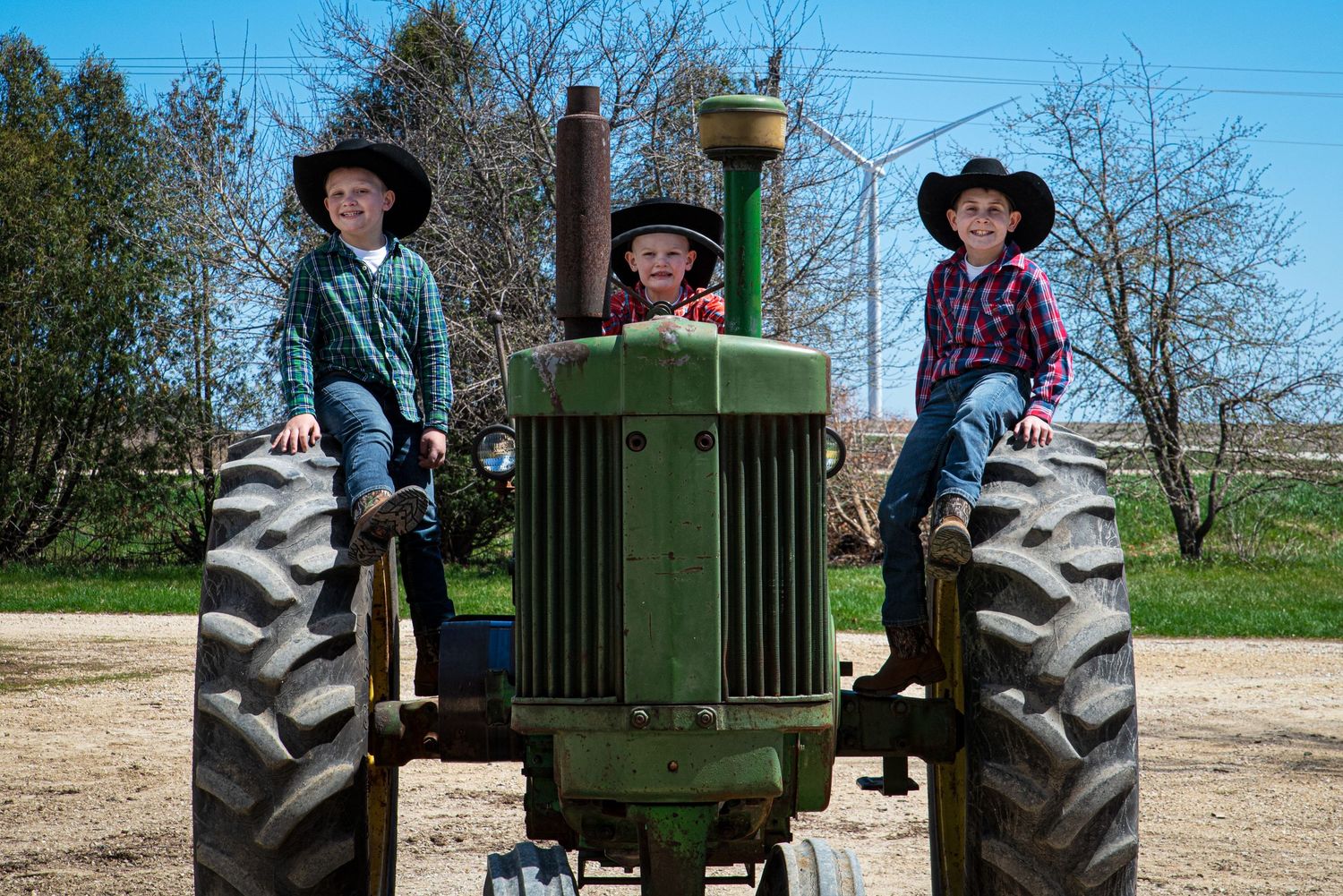 Three children in cowboy hats posing on a vintage green John Deere tractor on a sunny farm day.