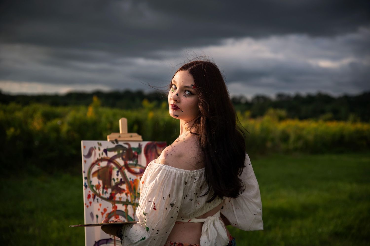 Young woman in white off-shoulder top painting on canvas outdoors under dramatic sky.