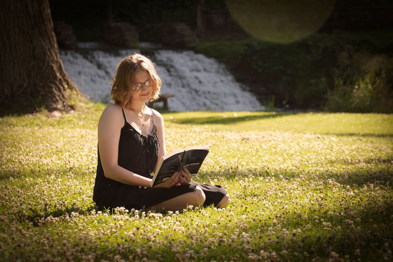 Blonde woman in black dress reading a book while sitting on a clover-covered lawn.