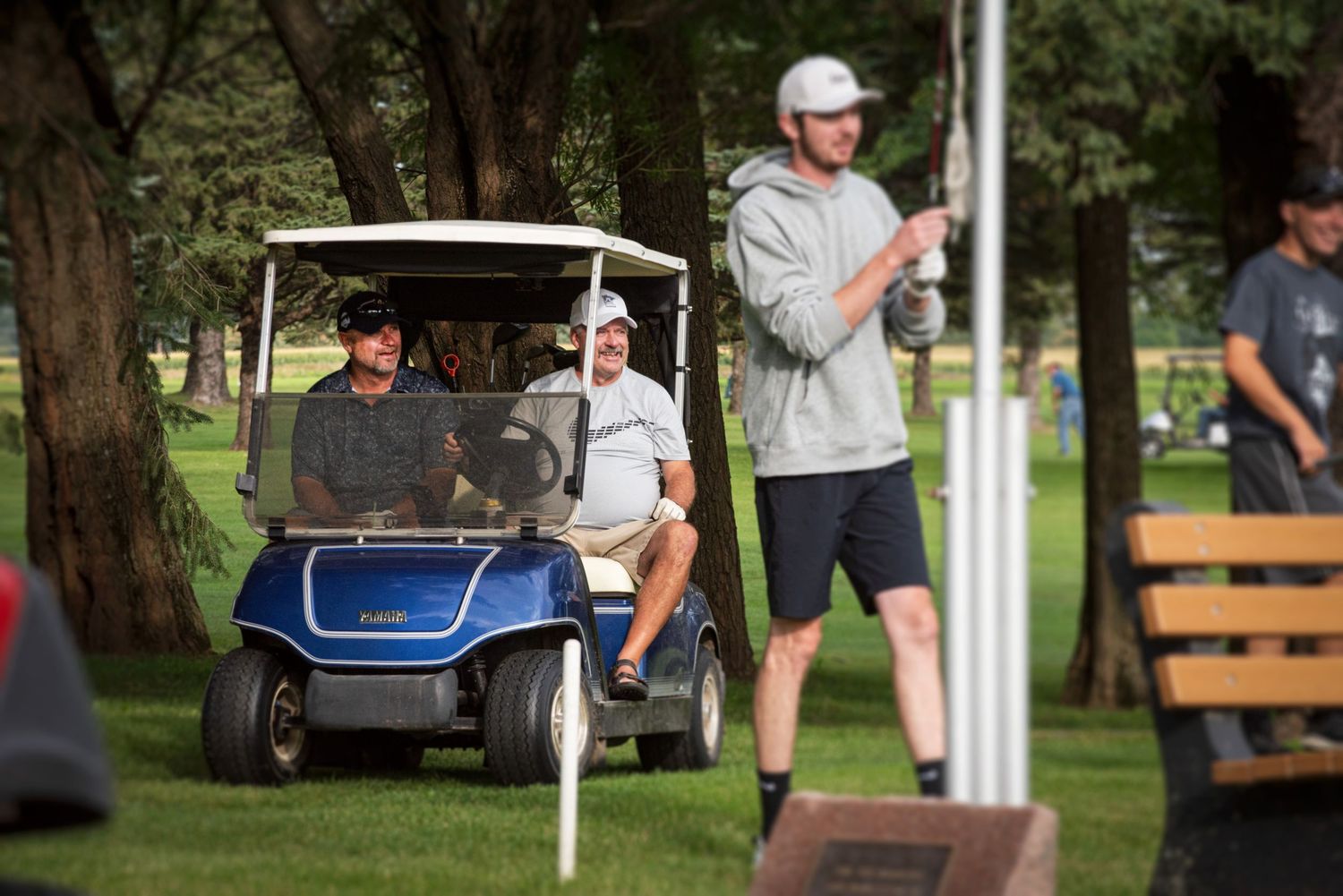 Golfers riding a blue golf cart on a tree-lined course during a sunny day.