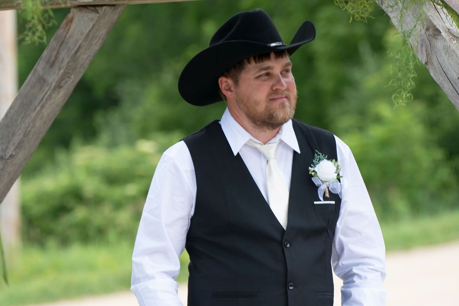 Groom in black cowboy hat and vest with boutonniere stands under wooden arch at outdoor wedding ceremony.