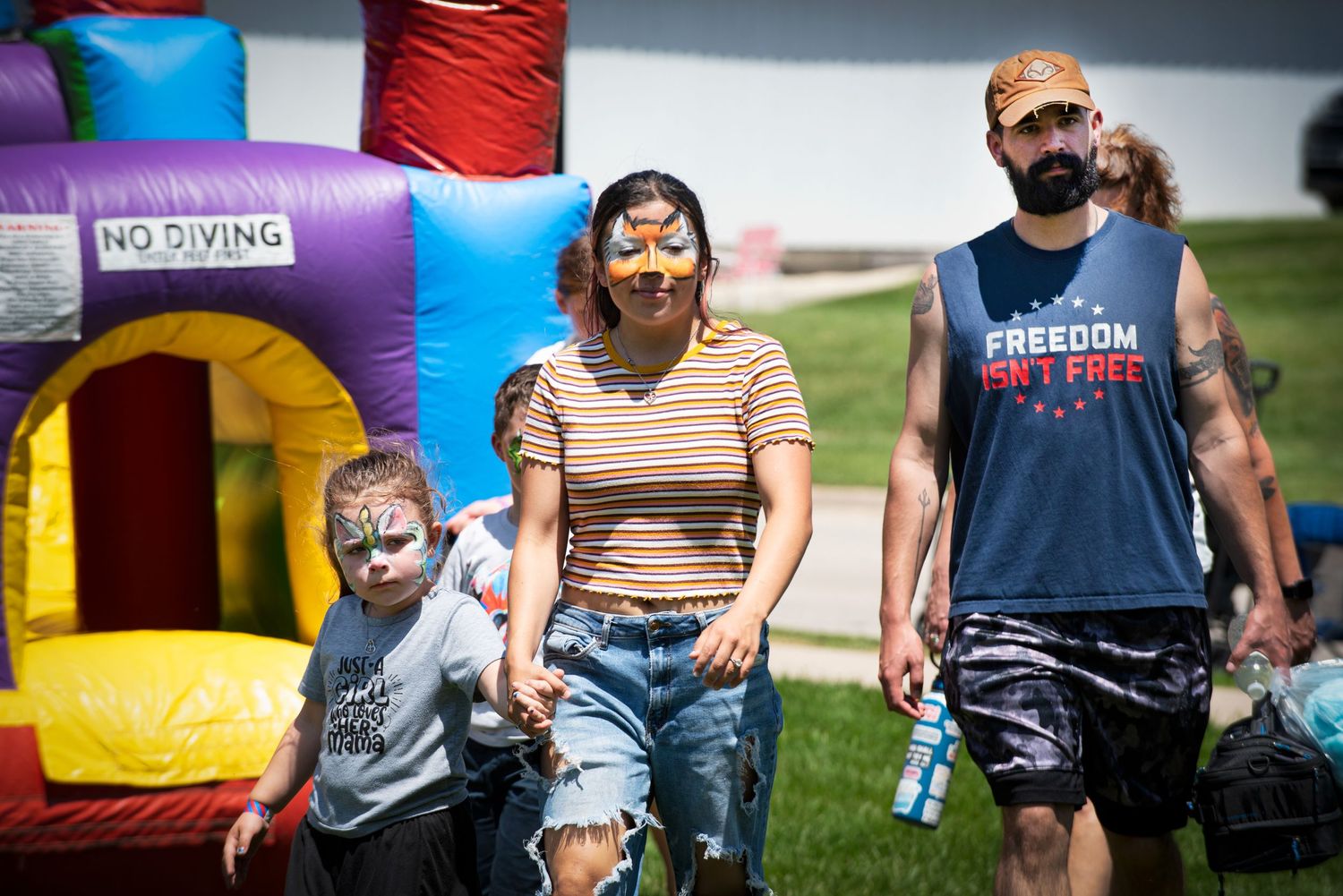 A family with face-painted children walks past a bounce house at an outdoor summer festival. At Harmony MN 4th of July Celebration.