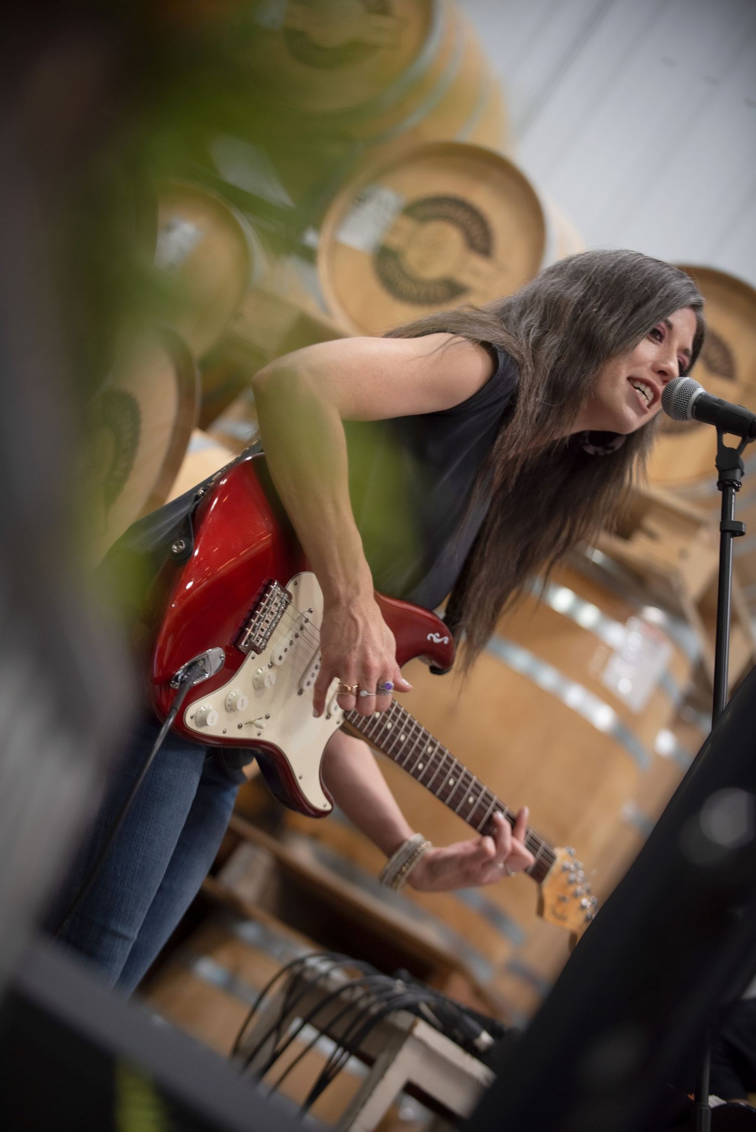 Female guitarist with long hair playing a red electric guitar and singing into a microphone at a distillery at Harmony Spirits..