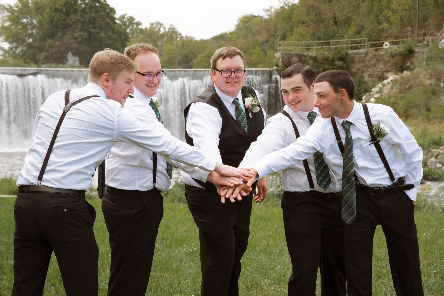 Groom and four groomsmen in suspenders stack hands together near waterfall in team celebration pose in Lanesboro MN.
