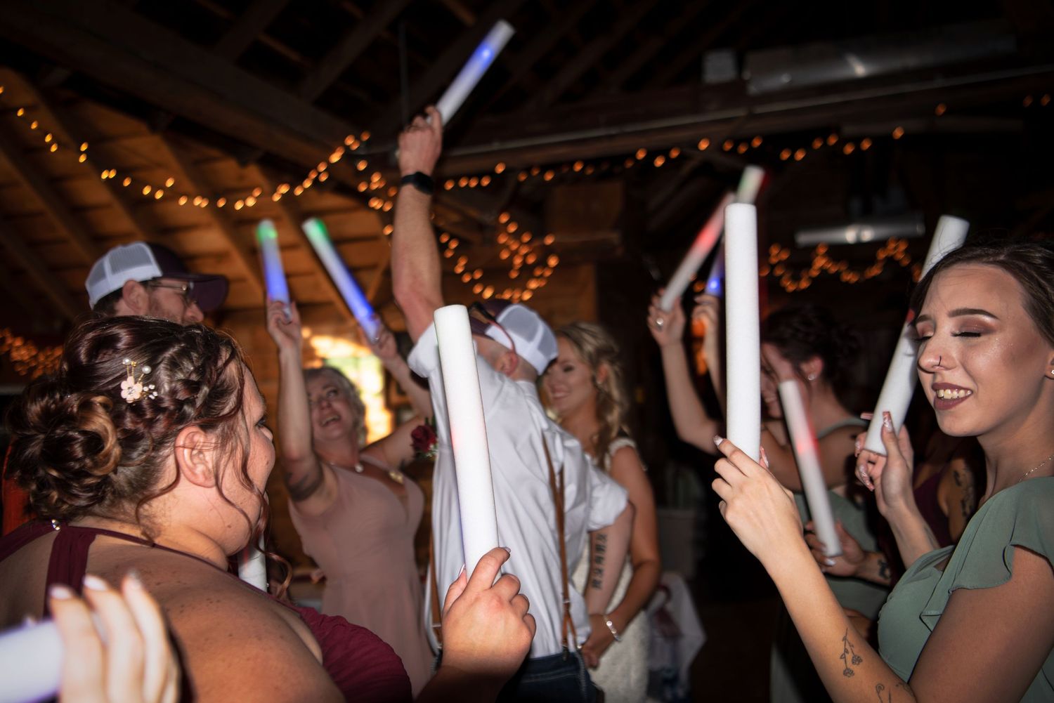 Wedding guests celebrate on dance floor holding glowing foam sticks under string lights at reception.