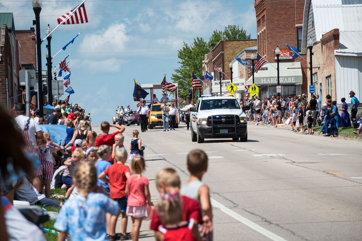 Crowds line a small-town Main Street to watch a patriotic Fourth of July parade with American flags and vehicles. At Harmony Mn.