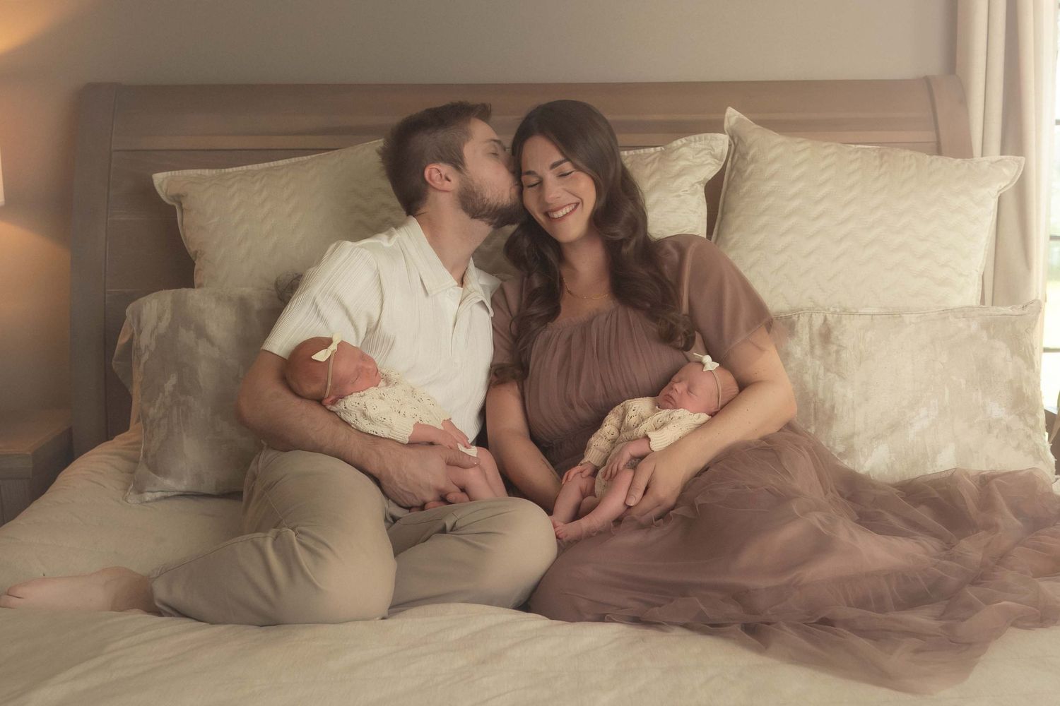 Parents sitting on a bed smiling at each other while each holding a newborn baby in soft warm lighting.