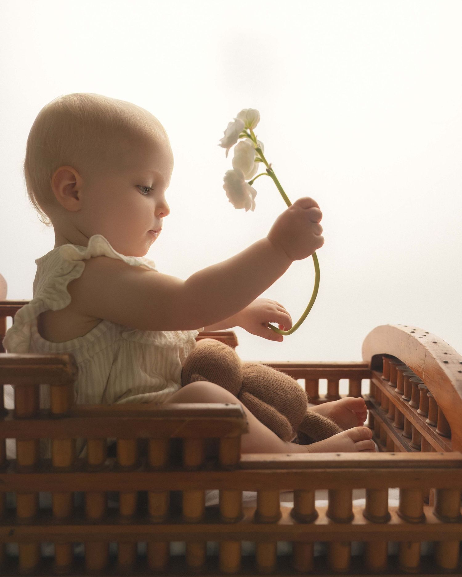 A baby girl in a white romper sits in a wooden crib, delicately holding a small white flower.