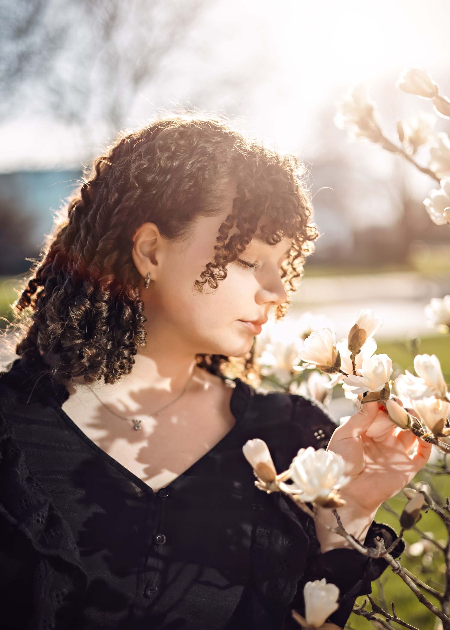 Young woman with curly hair smelling white magnolia blossoms in golden sunlight outdoors.