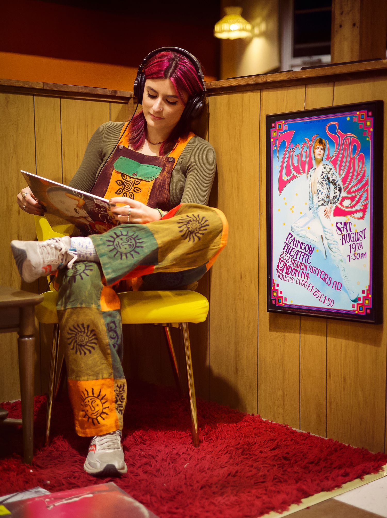 Woman with pink hair wearing headphones and colorful sun-print pants reads a vinyl record sleeve in a retro room.