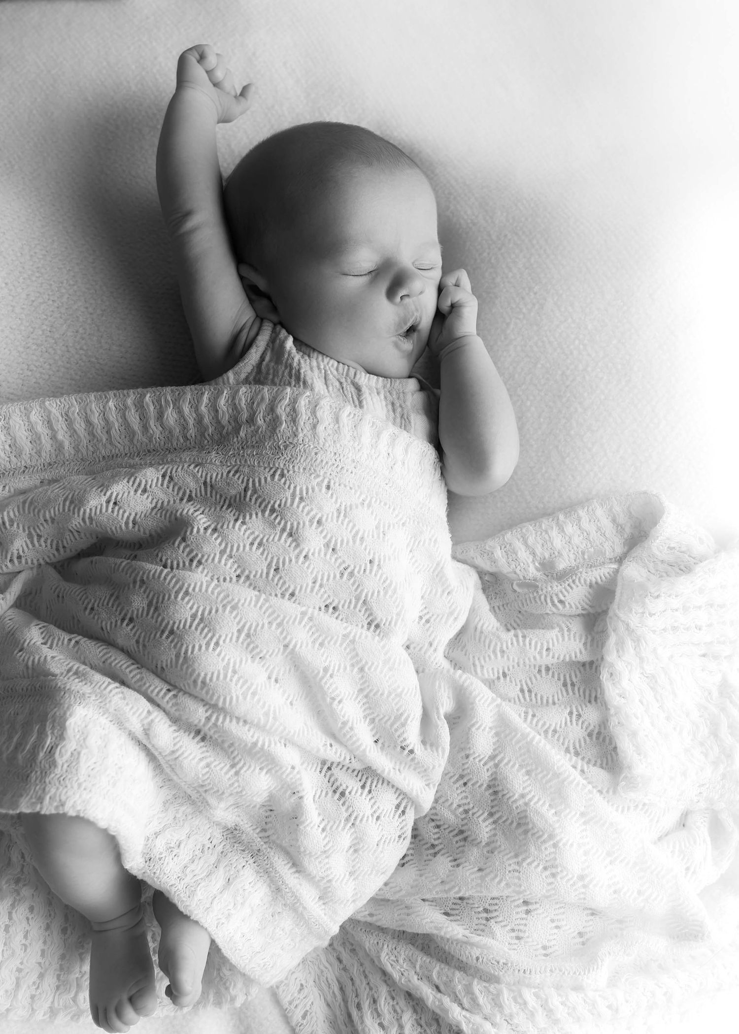 Black and white photo of a sleeping newborn baby in a white knit outfit with one arm raised.