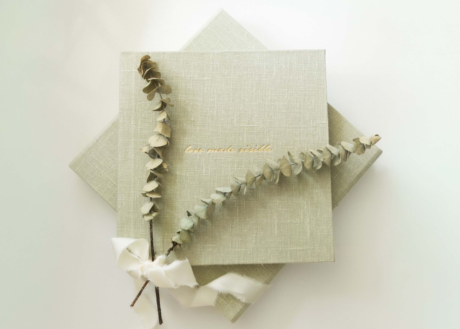 Elegant sage green linen photo album with gold foil text, adorned with a dried eucalyptus sprig on white background.
