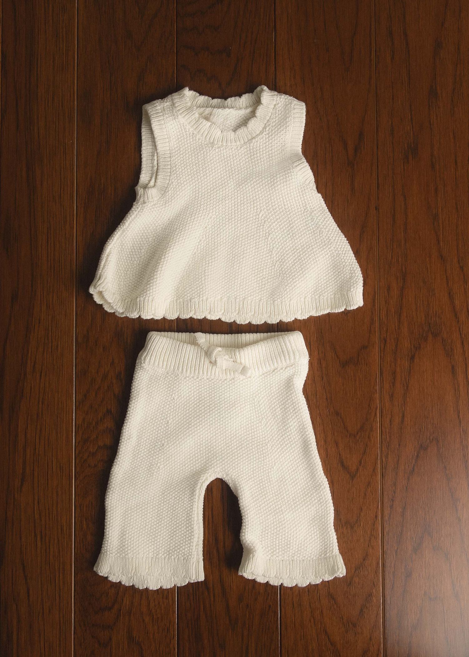 White knit baby two-piece outfit set with sleeveless top and pants with scalloped edges on wooden floor.