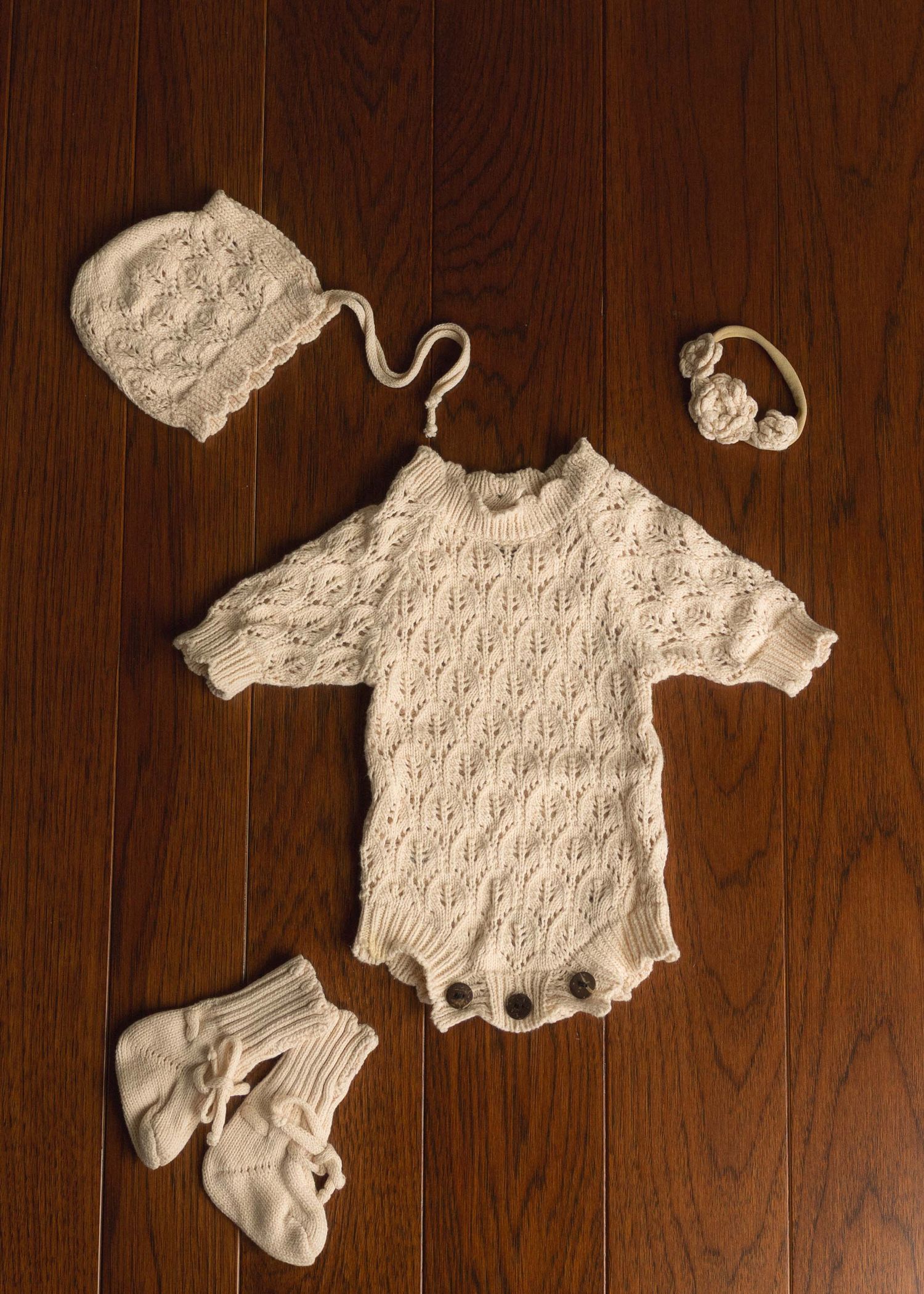 White knit baby romper set with matching bonnet, headband, and booties on wood floor.