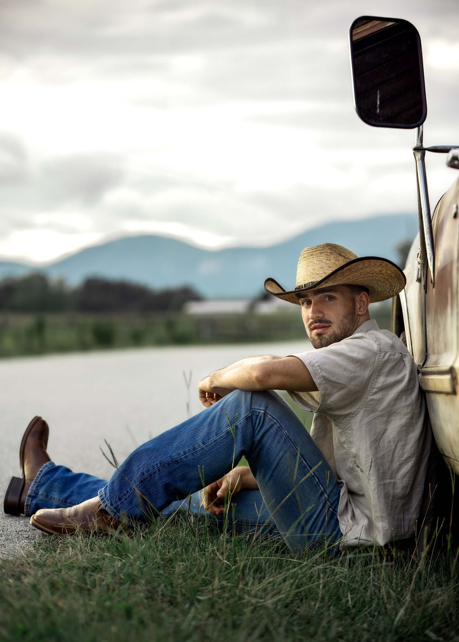 A young man in a straw cowboy hat, jeans, and boots sits against a vintage truck in a rural landscape.