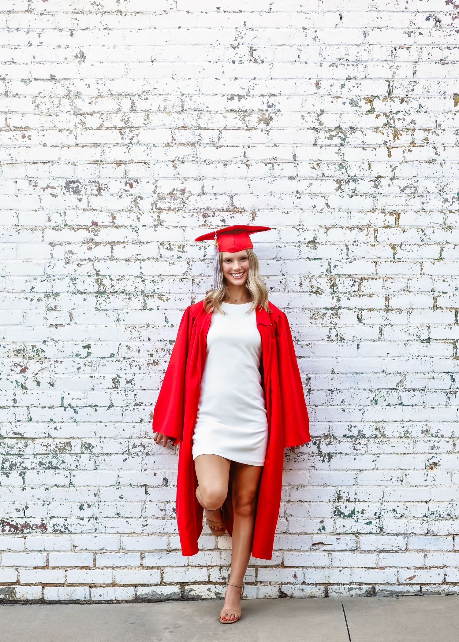 Smiling graduate in red cap and gown poses against white brick wall wearing a white dress and heels.