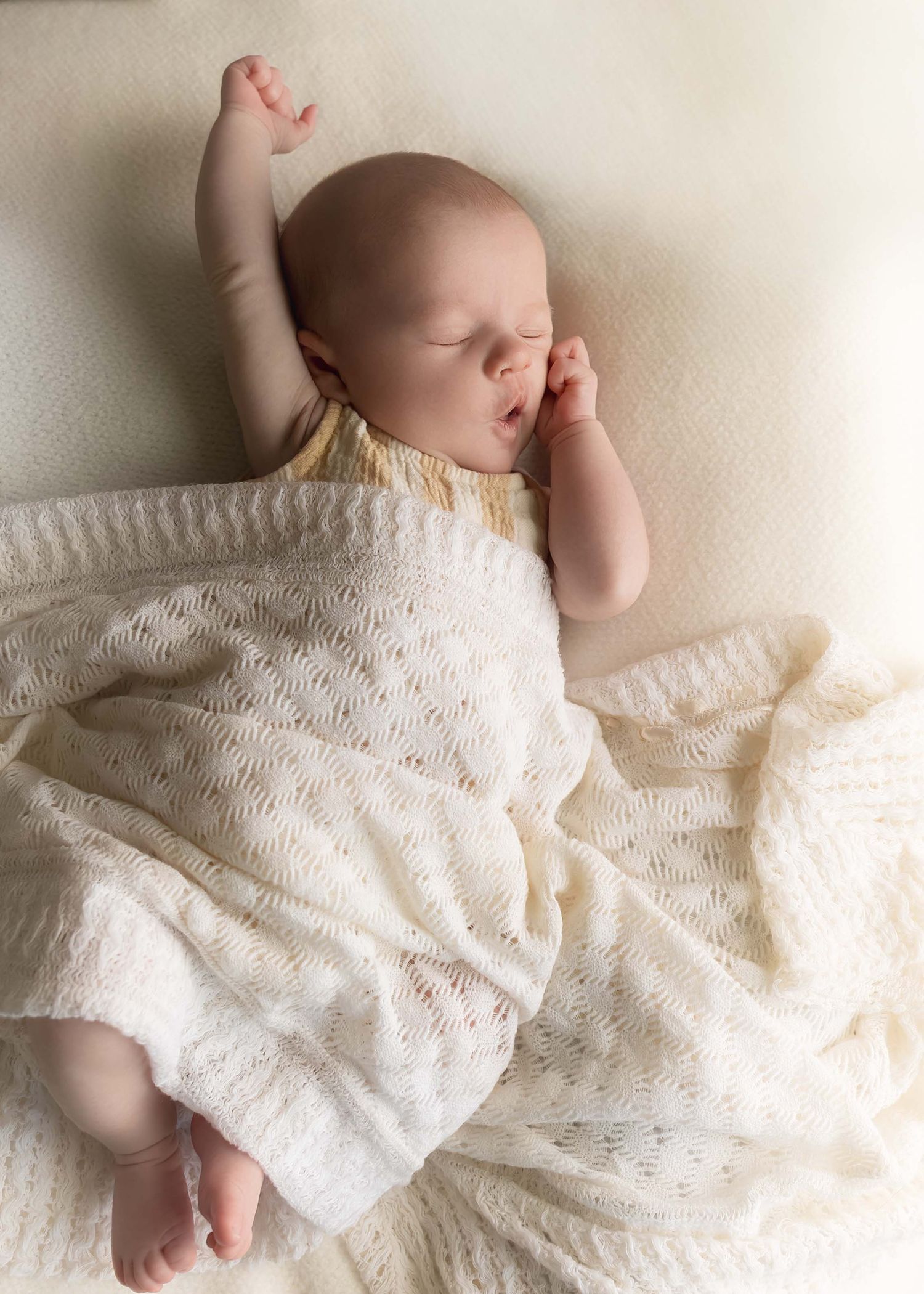Sleeping newborn baby in white knit blanket and yellow outfit with arm raised on cream background.
