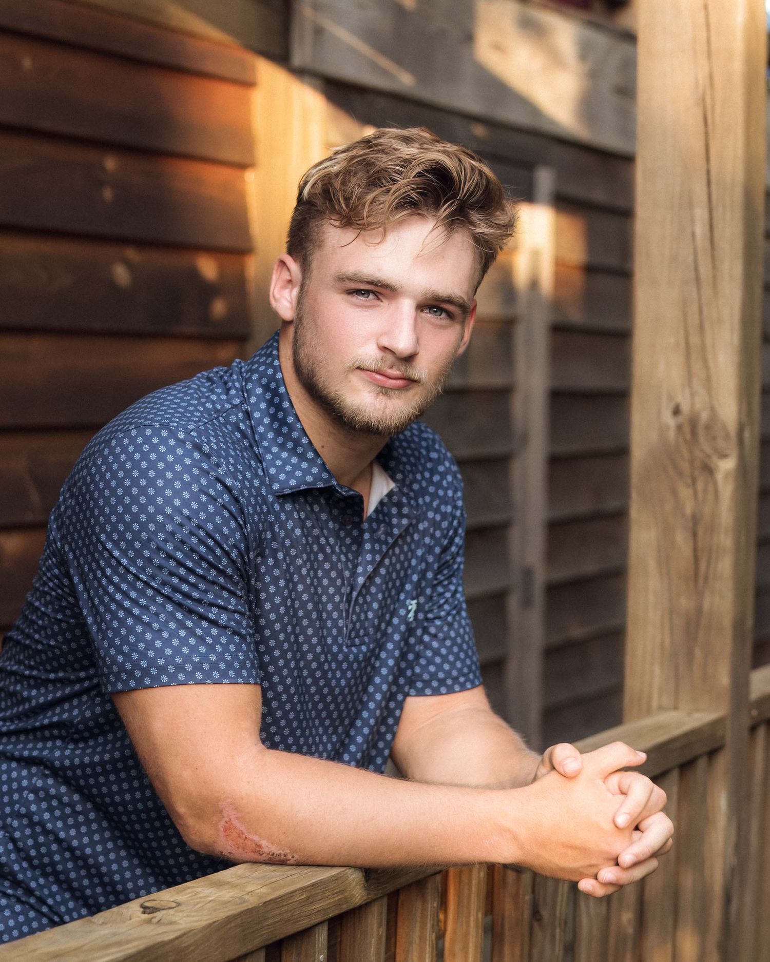 Young man in navy blue patterned shirt leaning on wooden fence outdoors in warm golden light.