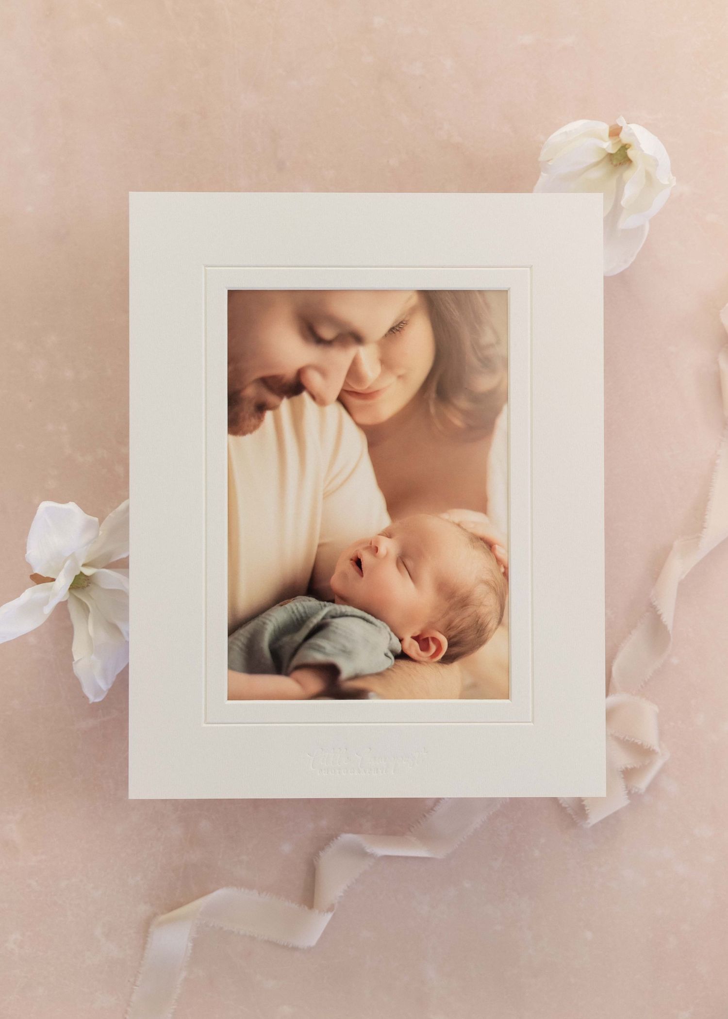 Framed newborn photo print showing parents lovingly gazing at their sleeping baby, displayed on a soft pink background.
