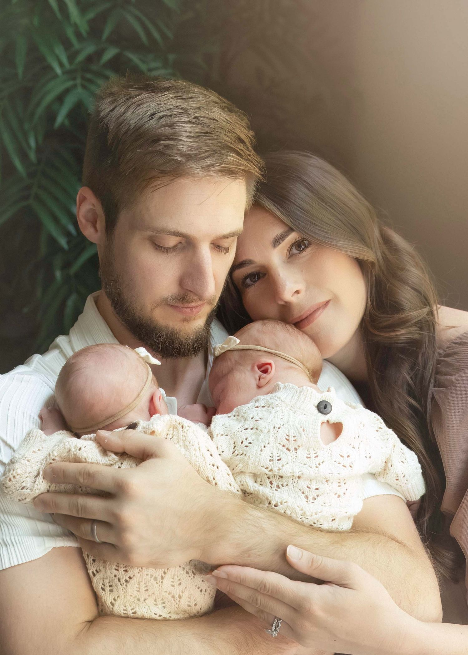 A couple tenderly holds their two newborn babies wrapped in white lace, posing for a family portrait near green foliage.