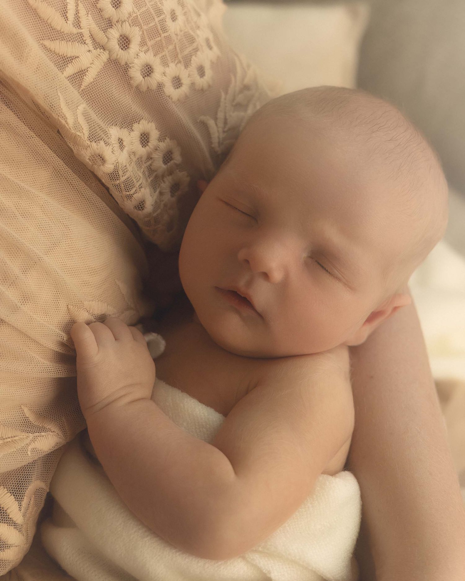 A peaceful newborn baby sleeping wrapped in a white cloth, cradled in an adult's arms wearing lace.