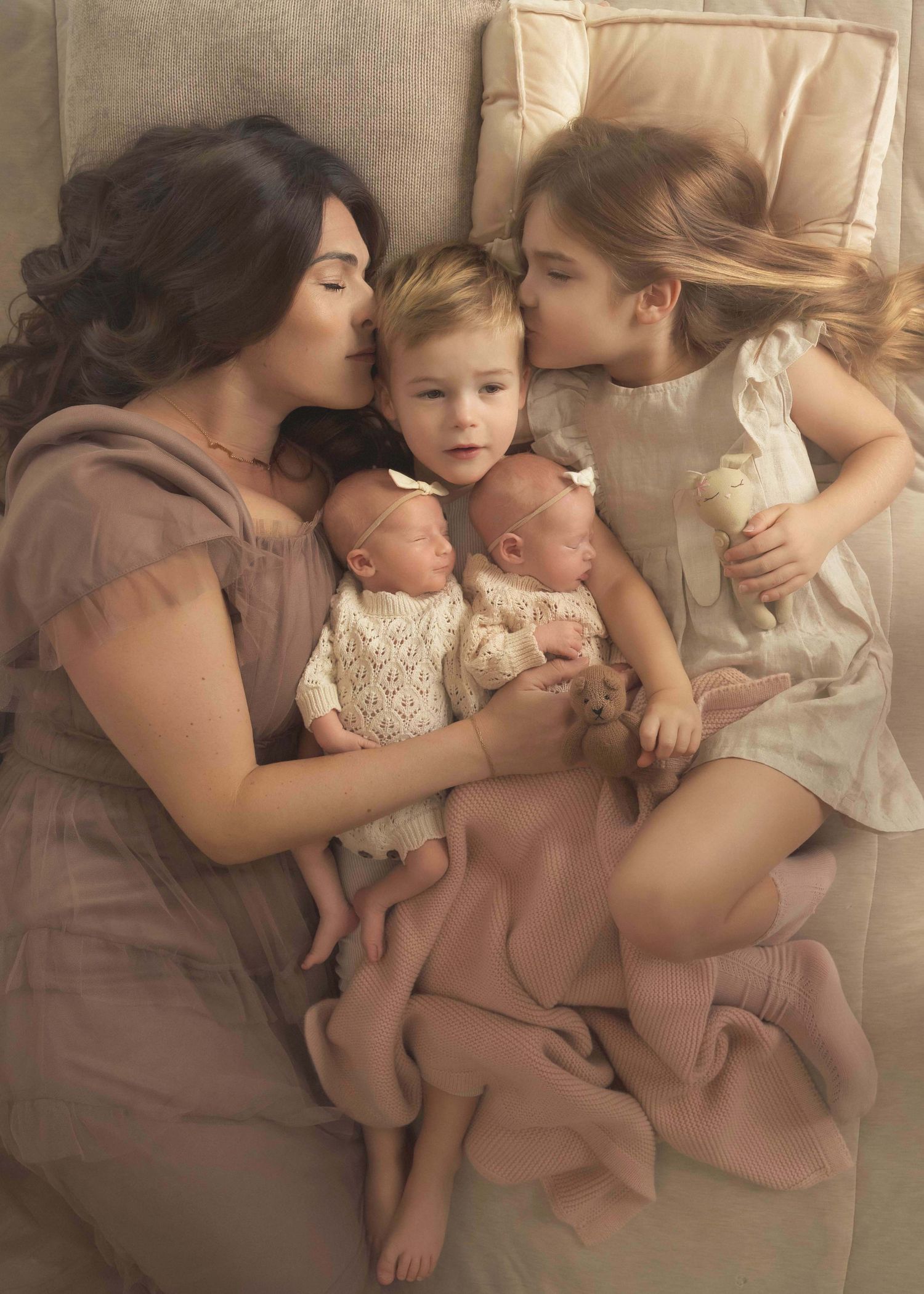 A mother lies with her four children, including newborn twins, as older siblings share tender kisses in a soft, dreamy portrait.