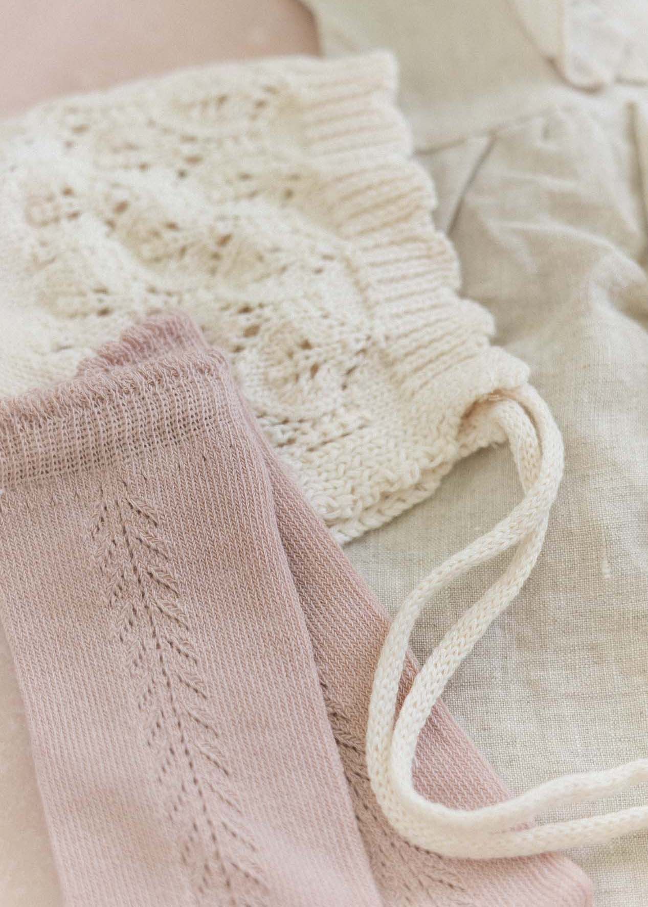 Soft knit baby clothing in cream and blush pink, featuring delicate lace and leaf patterns with ribbon ties.