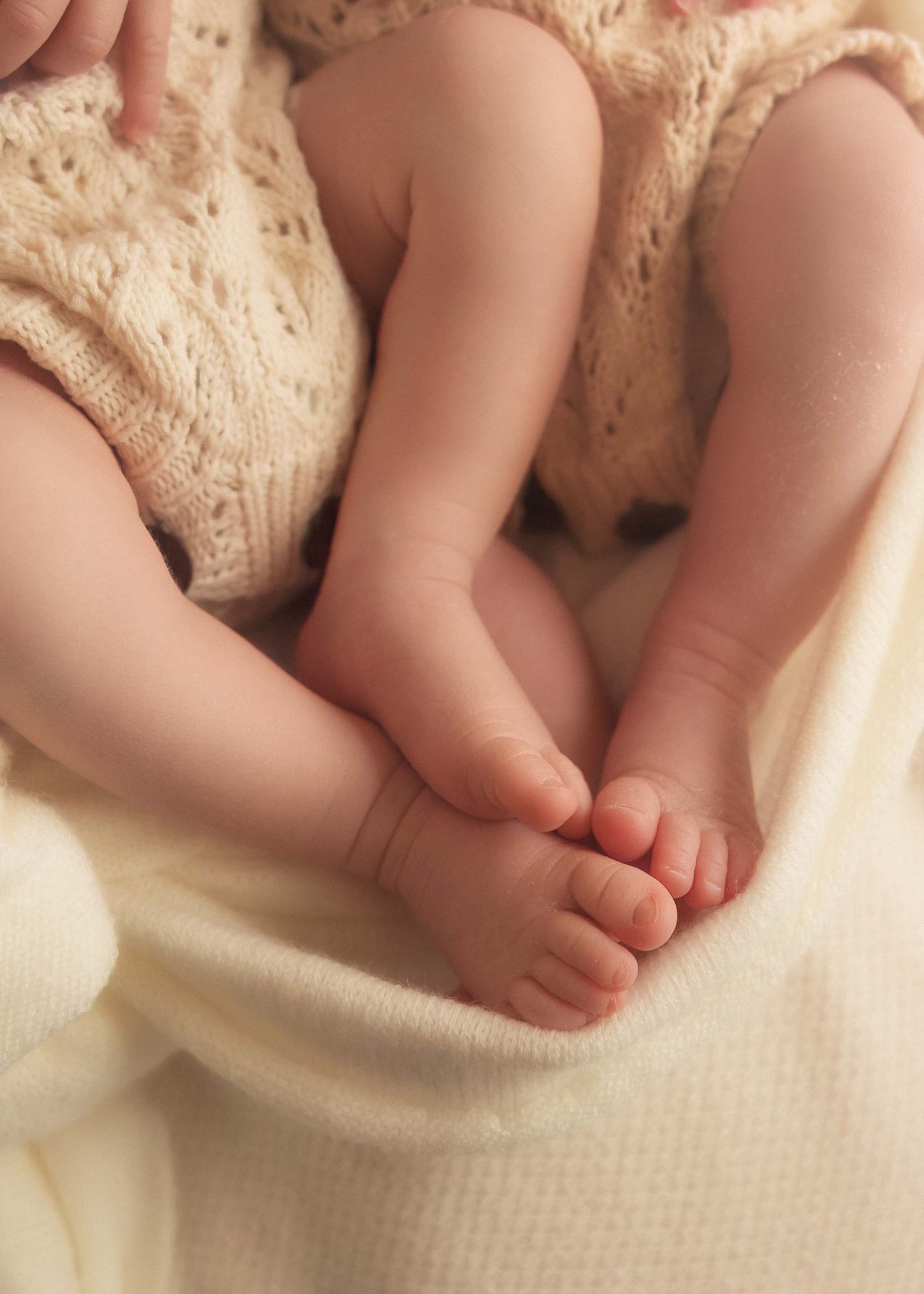 Close-up of two newborn babies' tiny feet and legs in cream knit rompers resting on a soft white blanket.