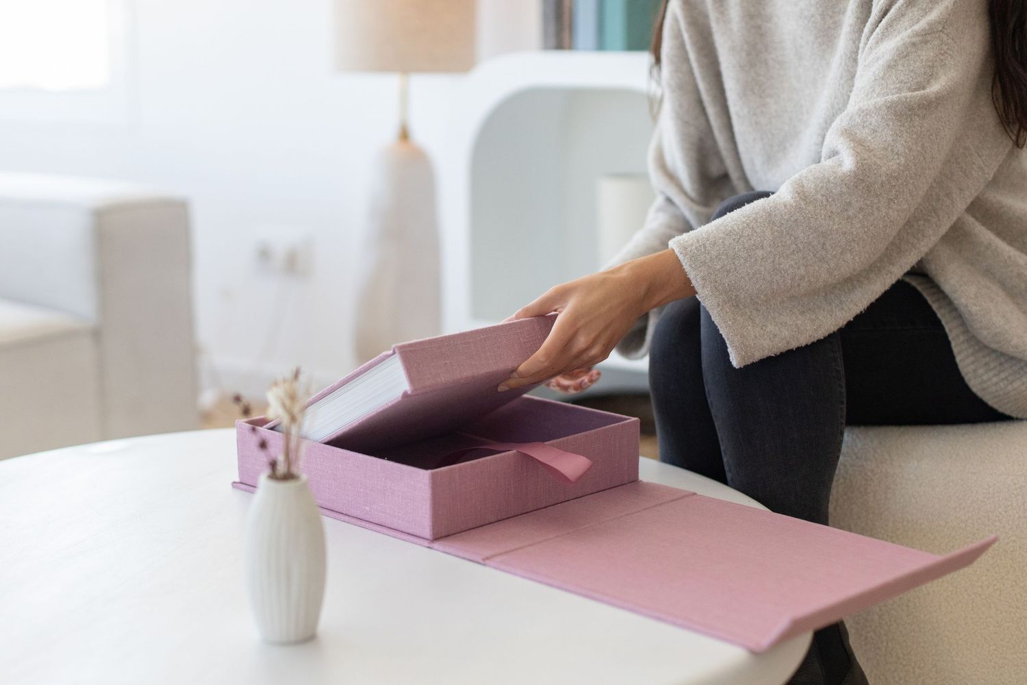 Woman in gray sweater opening a pink album box on a white table with a small vase nearby.