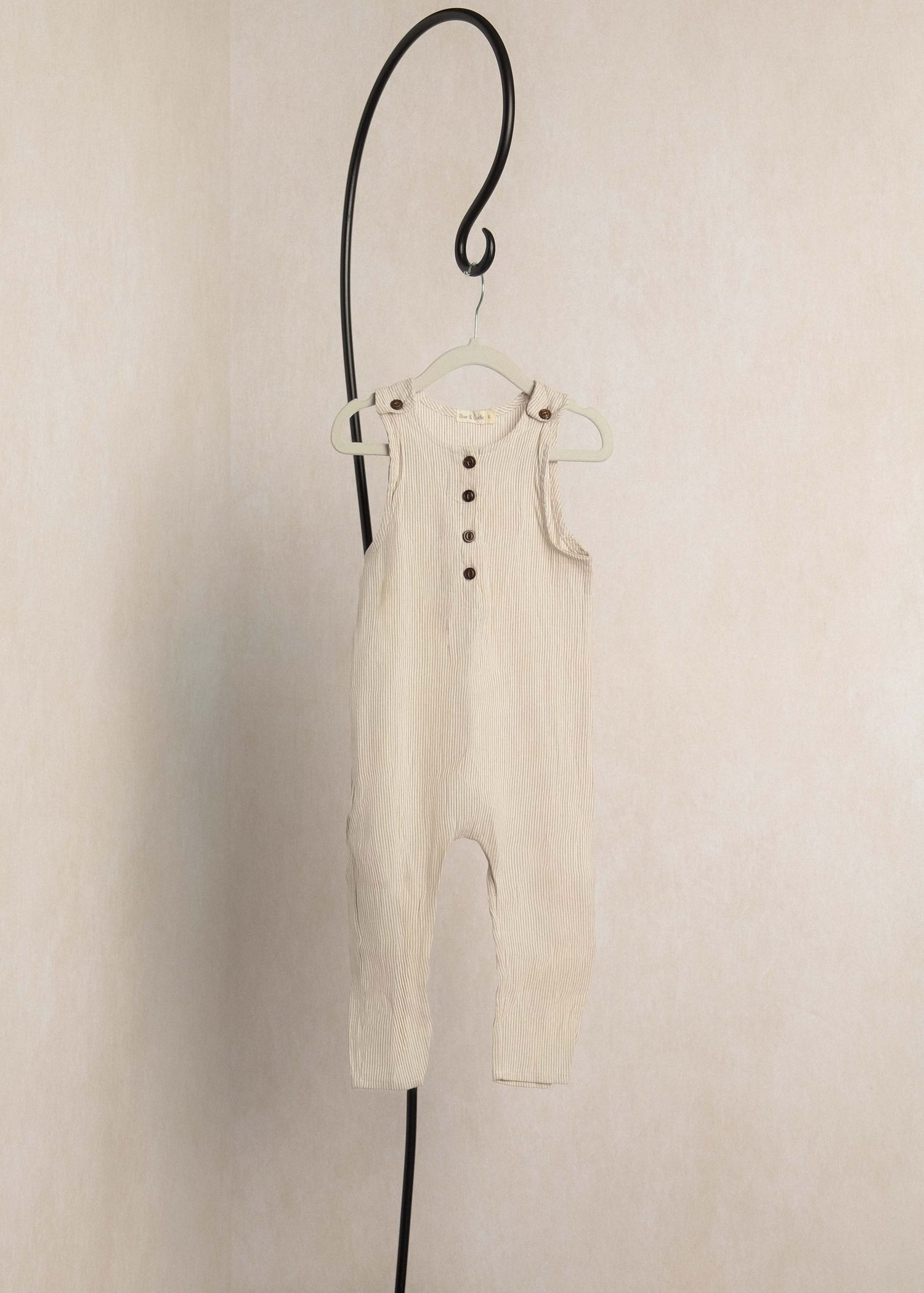Cream ribbed sleeveless baby romper with brown buttons, hanging on a decorative black hook against a neutral wall.