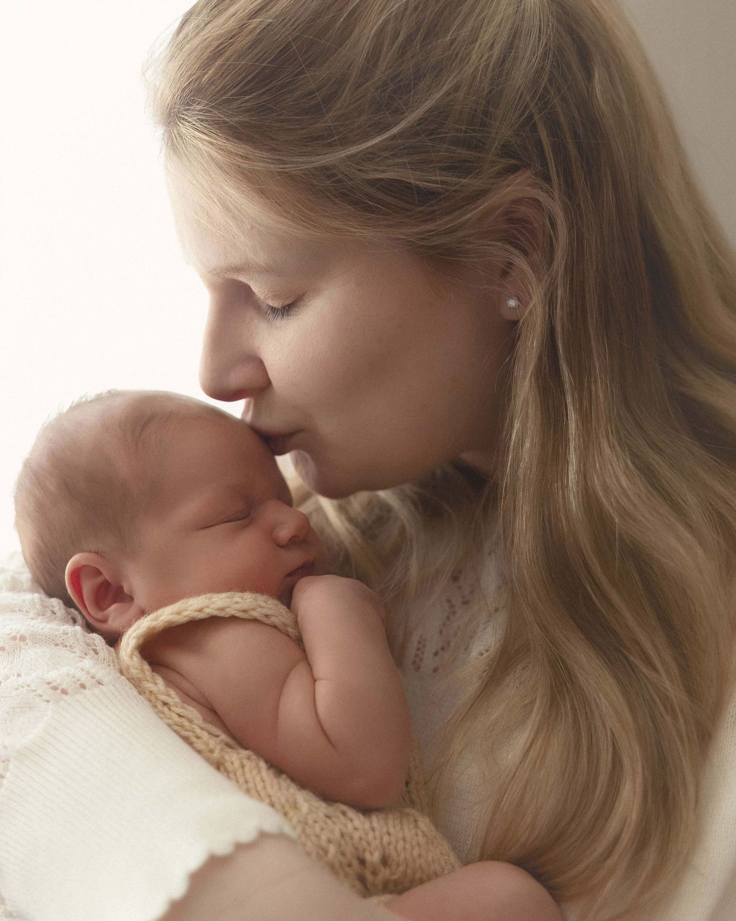 A tender moment as a young blonde mother gently kisses her sleeping newborn baby wrapped in a cream knit blanket.