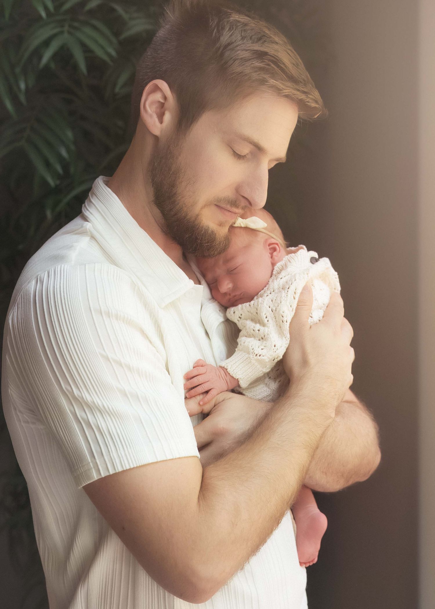 A bearded man in a white shirt tenderly holds a sleeping newborn baby close to his face.