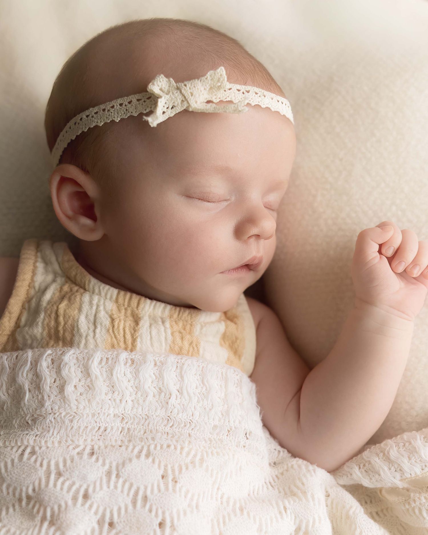 Sleeping newborn baby girl wearing a delicate white lace headband with bow, wrapped in a soft knit blanket.