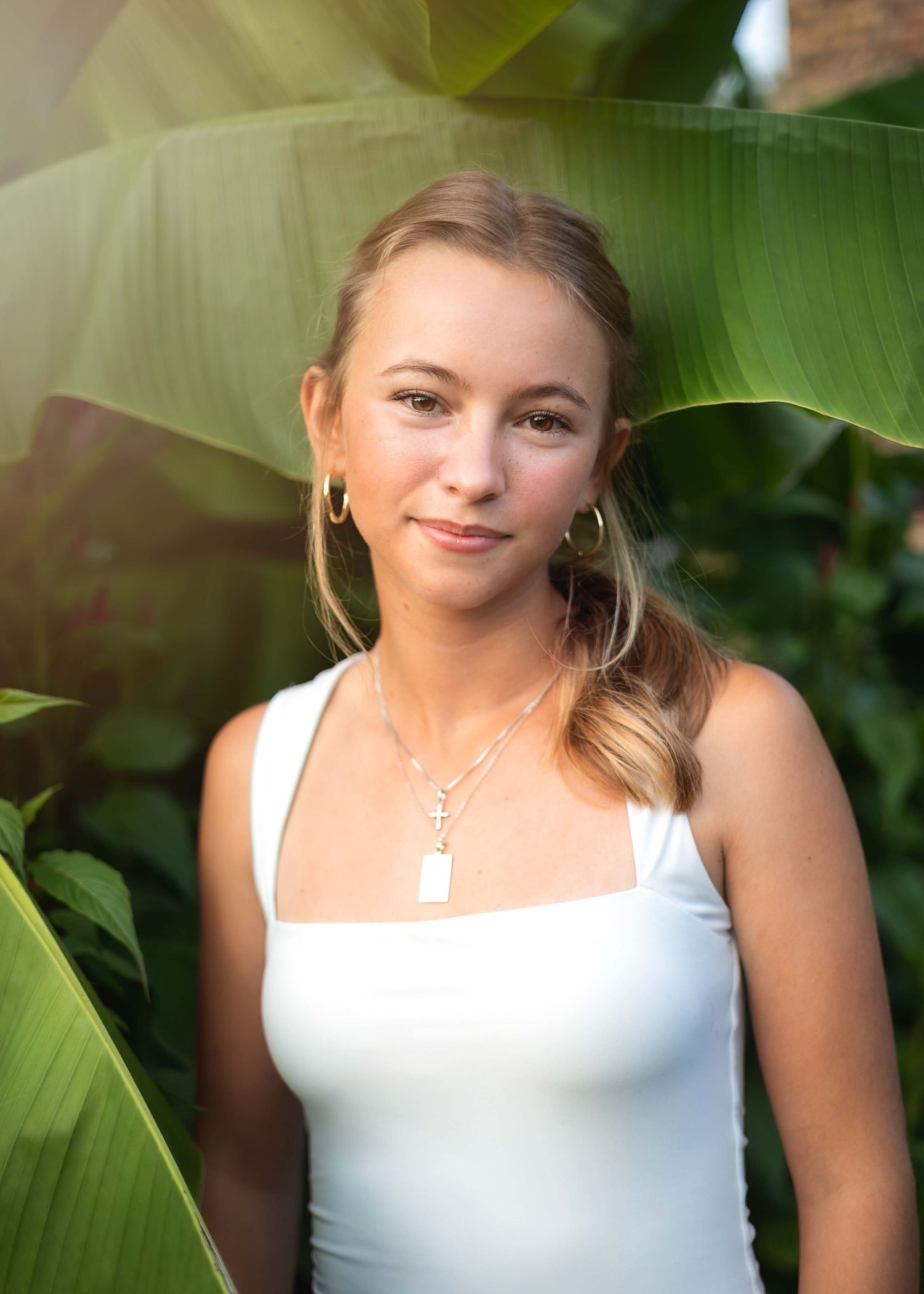 Young woman in white tank top with layered necklaces poses among tropical green leaves in warm natural light.