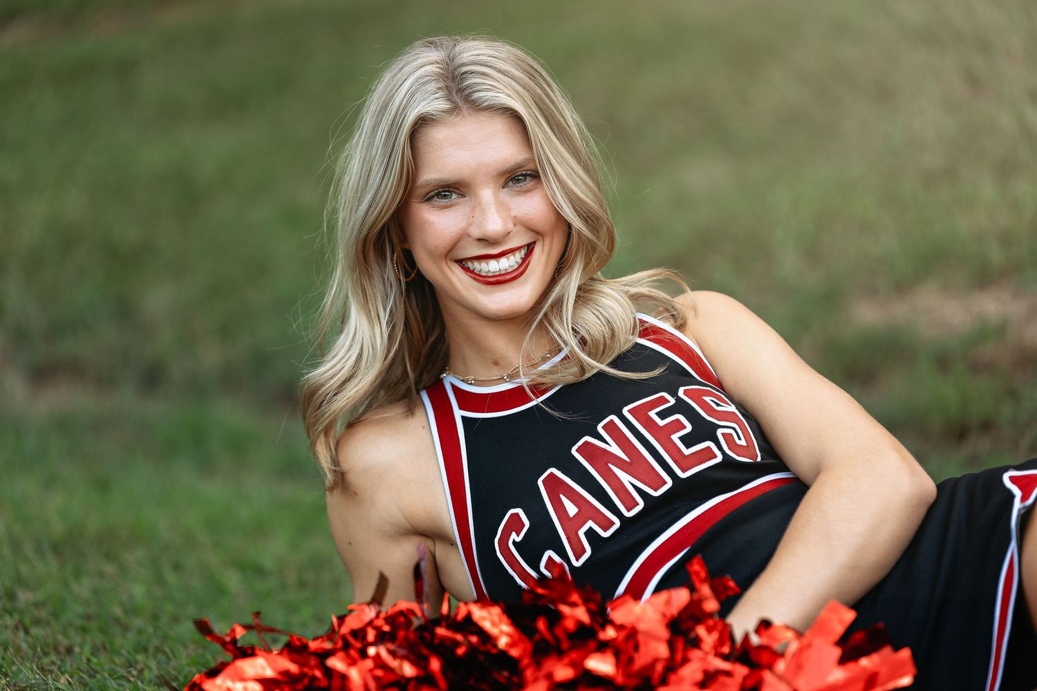 Smiling cheerleader in black and red Canes uniform holding red pom-poms outdoors on green grass.