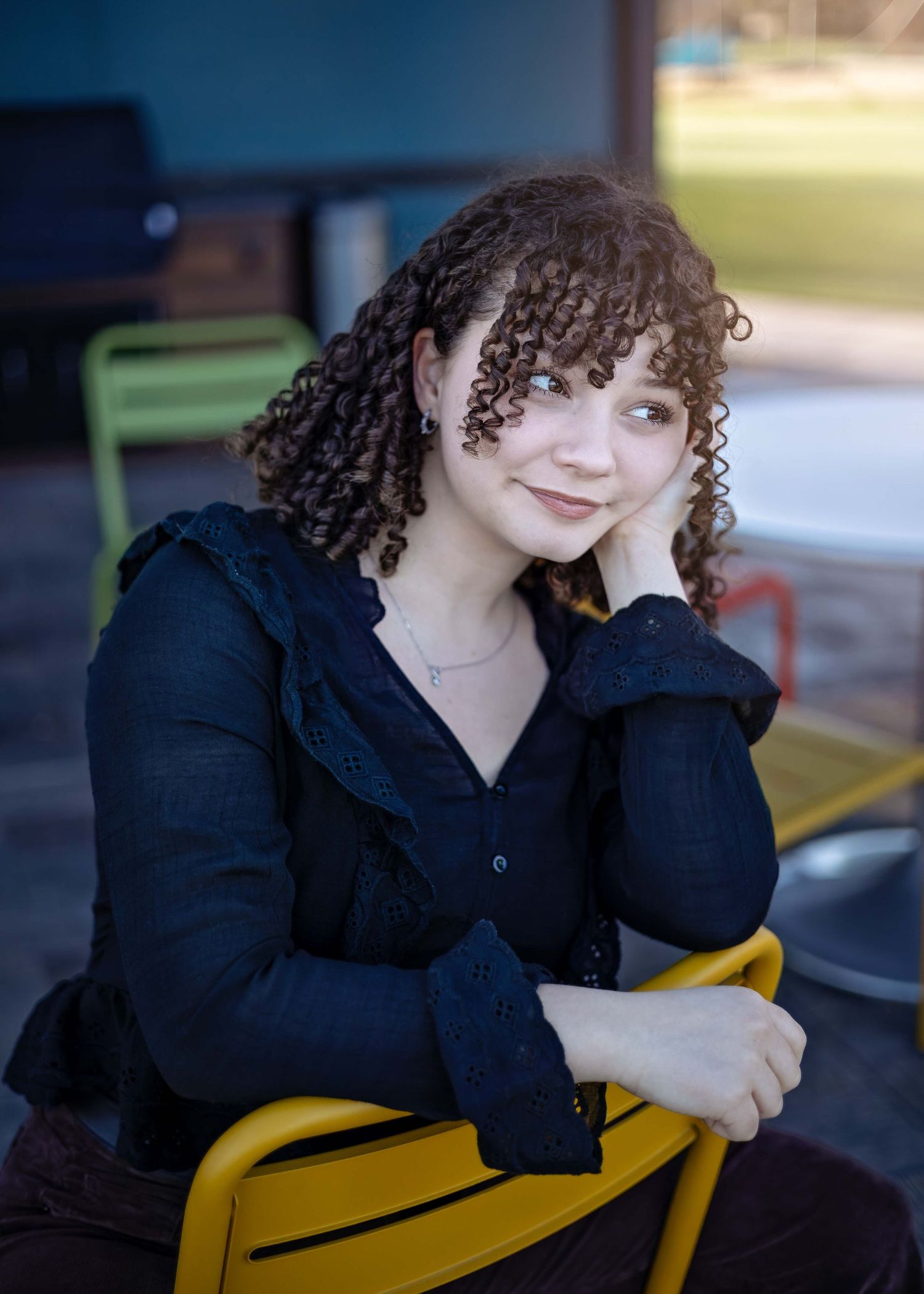 Young woman with curly hair sits relaxed on a yellow chair, wearing a black blouse, resting her chin on her hand.