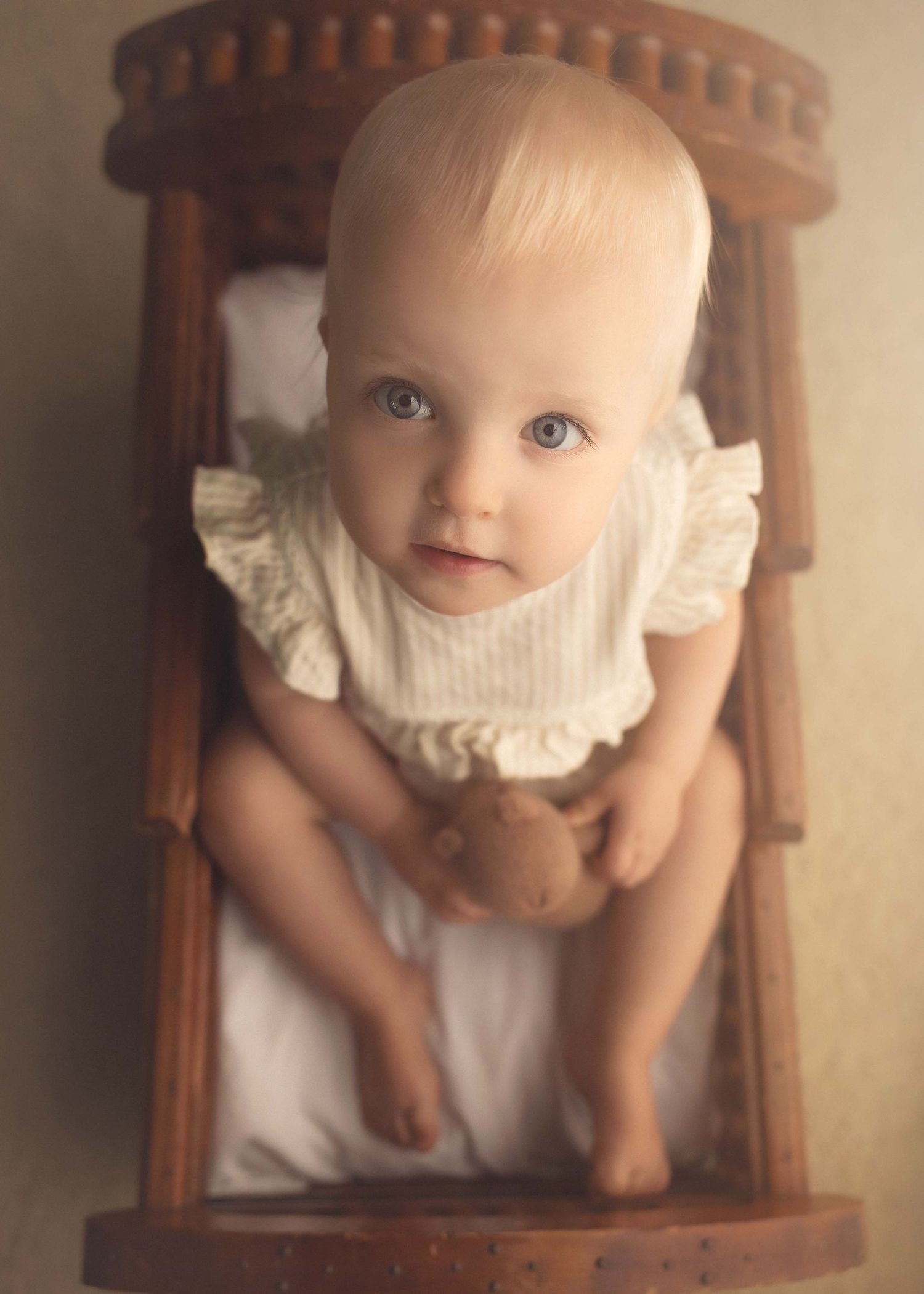 Adorable blue-eyed baby in white ruffle outfit sitting in a wooden chair holding a teddy bear.