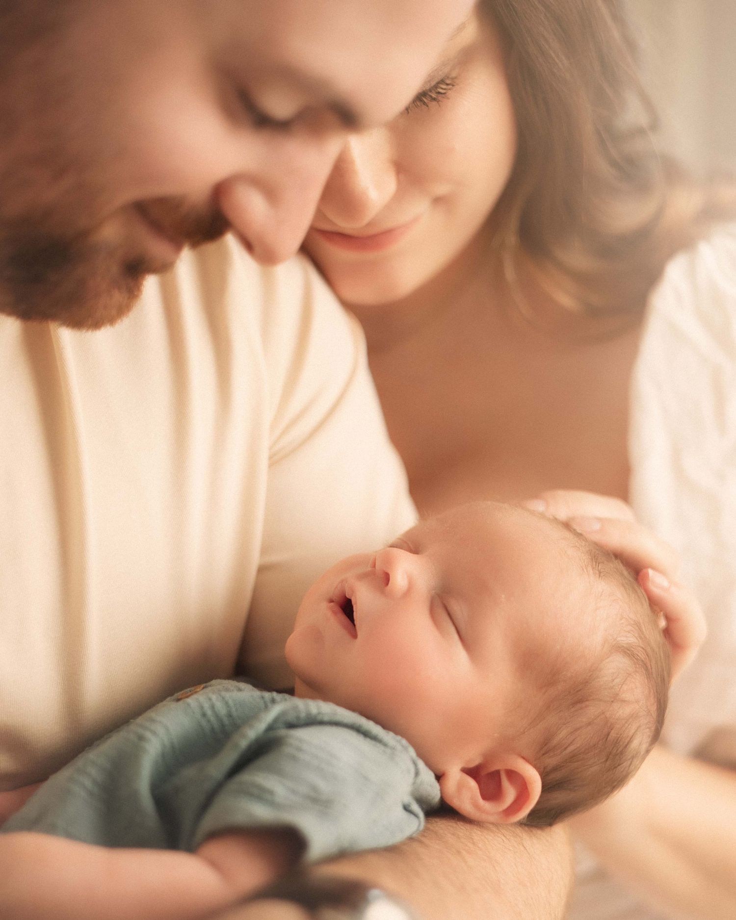 Two parents lovingly gazing at their newborn baby wrapped in a soft blue blanket in a warm, intimate portrait.