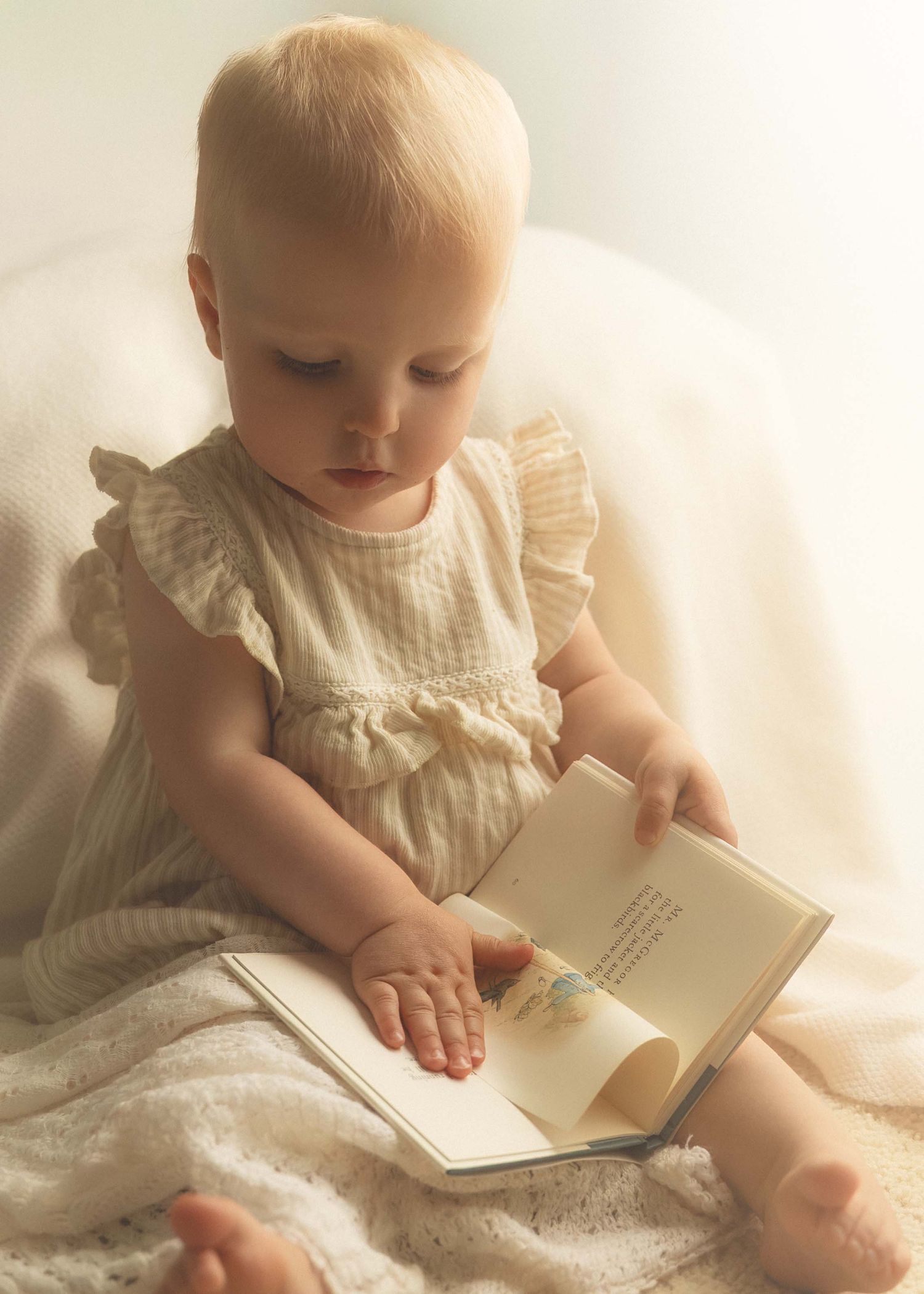 Adorable baby girl in a white ruffle dress sitting and reading a small book on a soft white blanket.