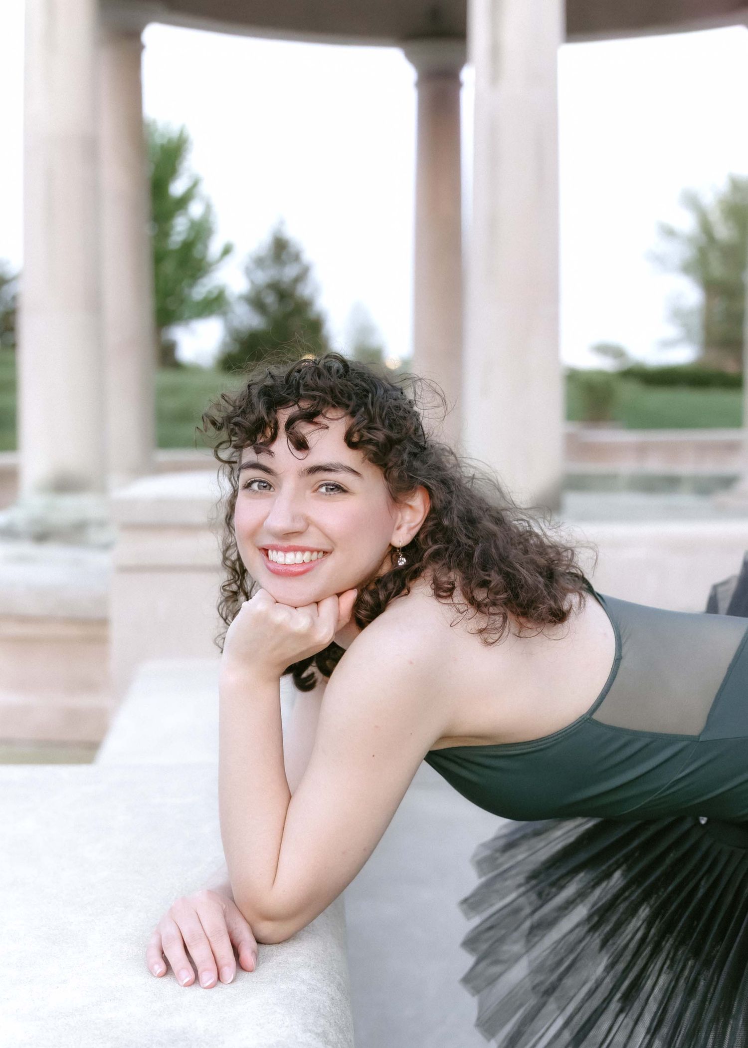 Young woman with curly hair smiles in elegant dark tulle dress, posing near classical columns outdoors.