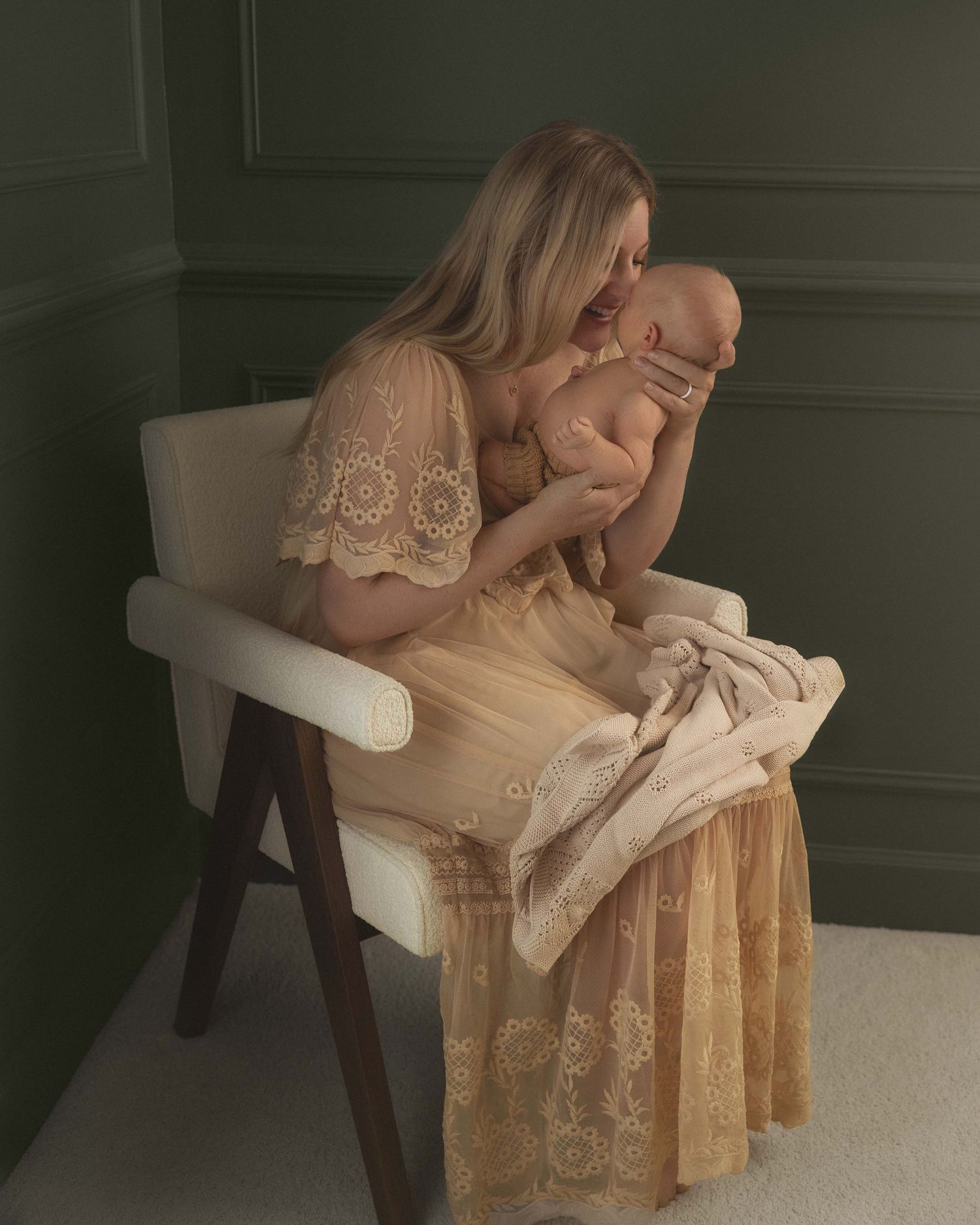 A mother in a lace gown holds her newborn baby while seated on a white chair in a soft, elegant studio setting.