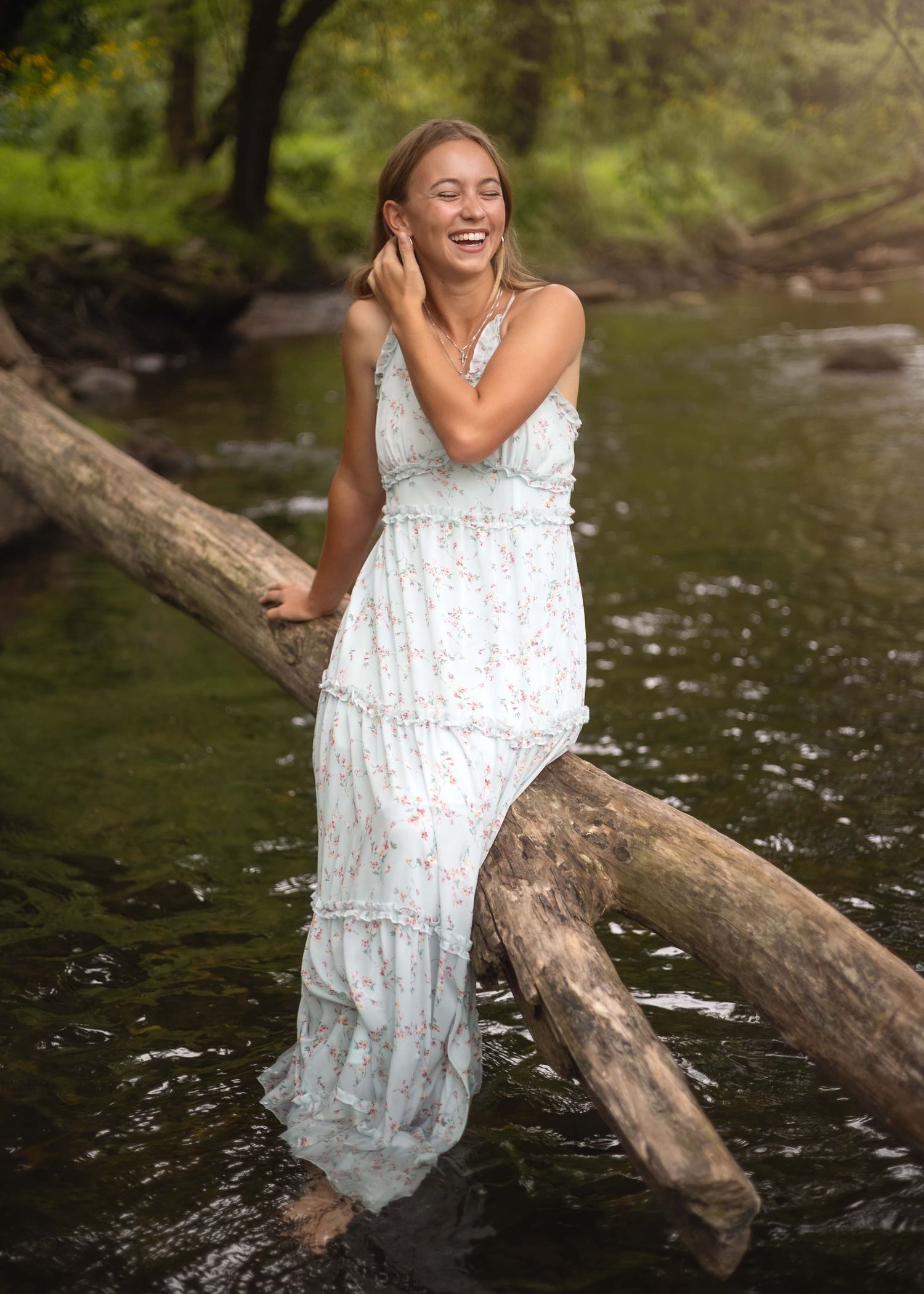 Young woman in floral maxi dress laughing by a forest creek, leaning on fallen logs in nature.