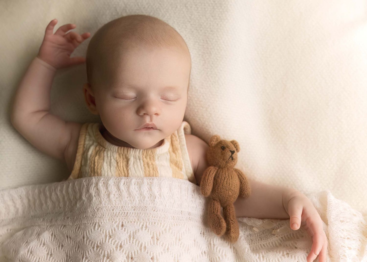Peaceful newborn baby sleeping with a small brown teddy bear on a cream knit blanket.