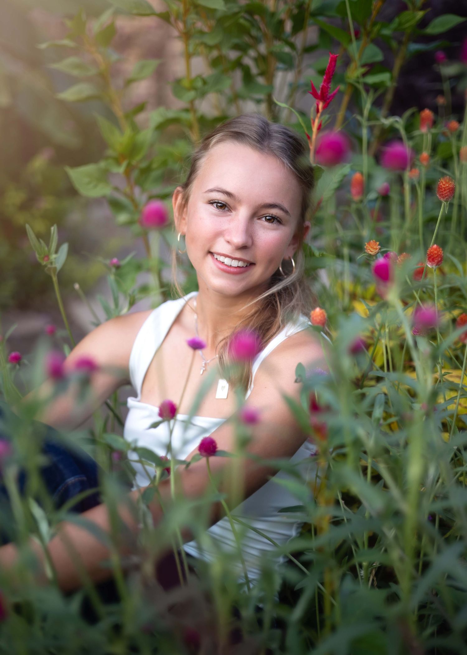 Young blonde woman smiling among colorful wildflowers in a lush garden, wearing a white top.